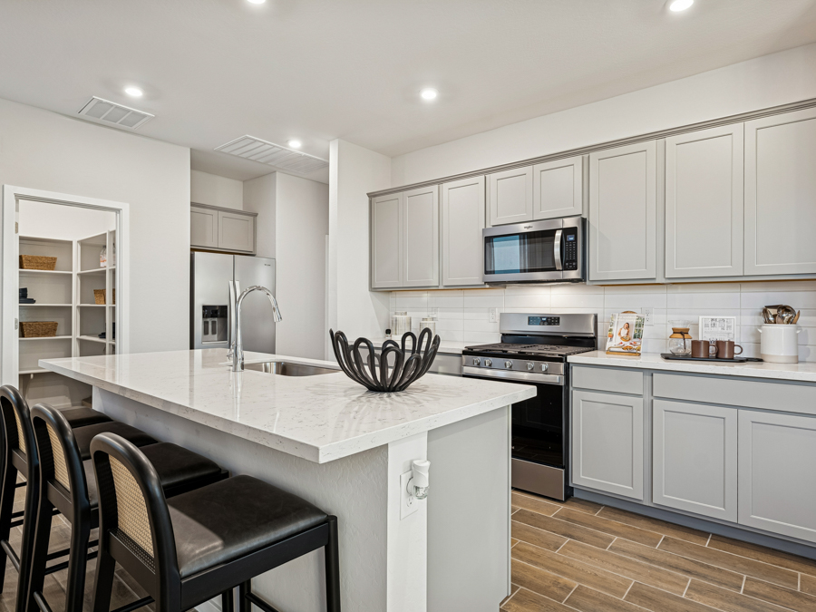 Modern kitchen with gray cabinets, stainless steel appliances, an island with a sink, three barstools, and a decorative bowl; a walk-in pantry is visible in the background.