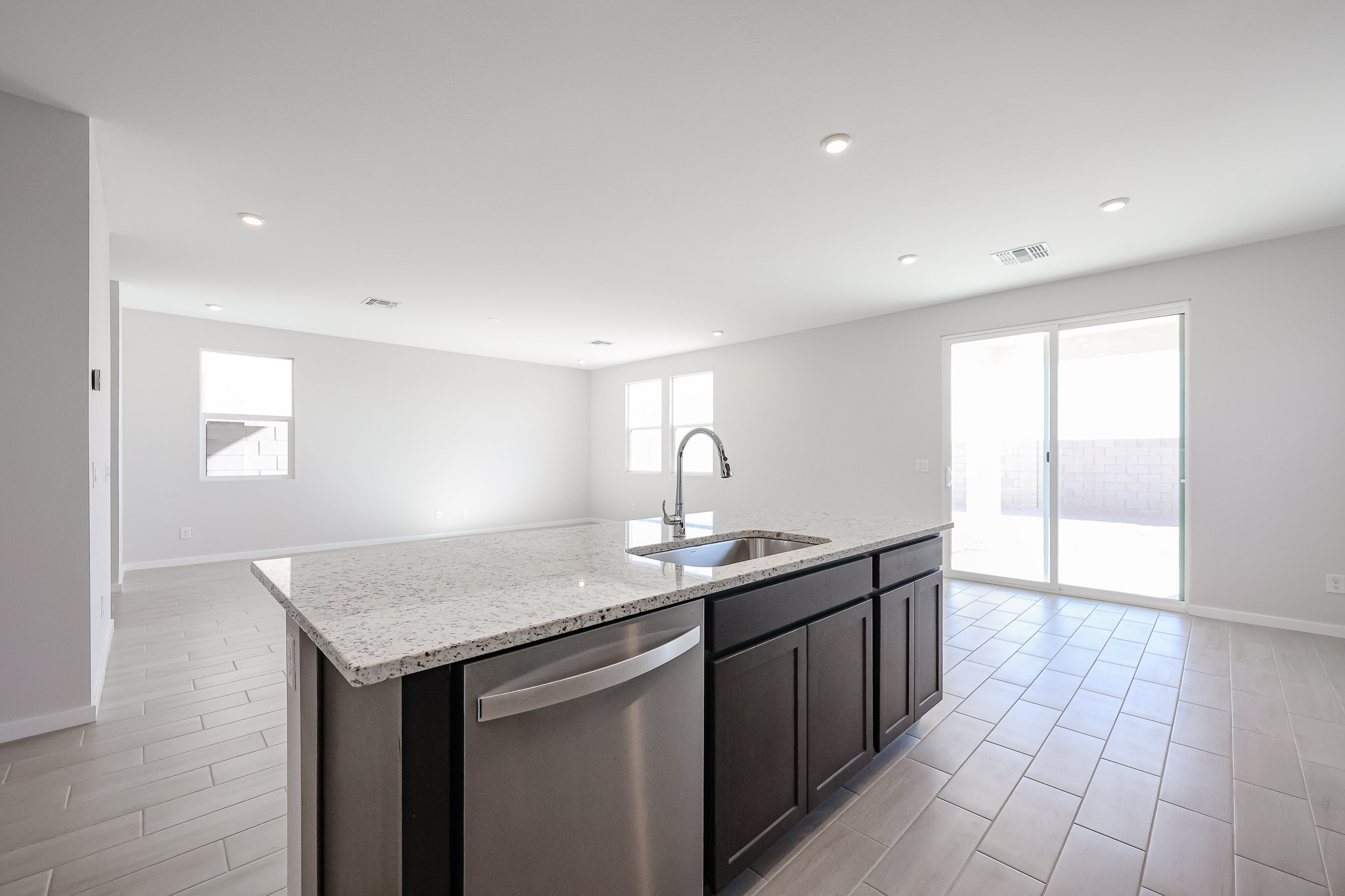 Modern, unfurnished kitchen with a granite island, dark cabinets, built-in sink, and stainless steel dishwasher, adjacent to a bright living area with tile floors and sliding glass doors.