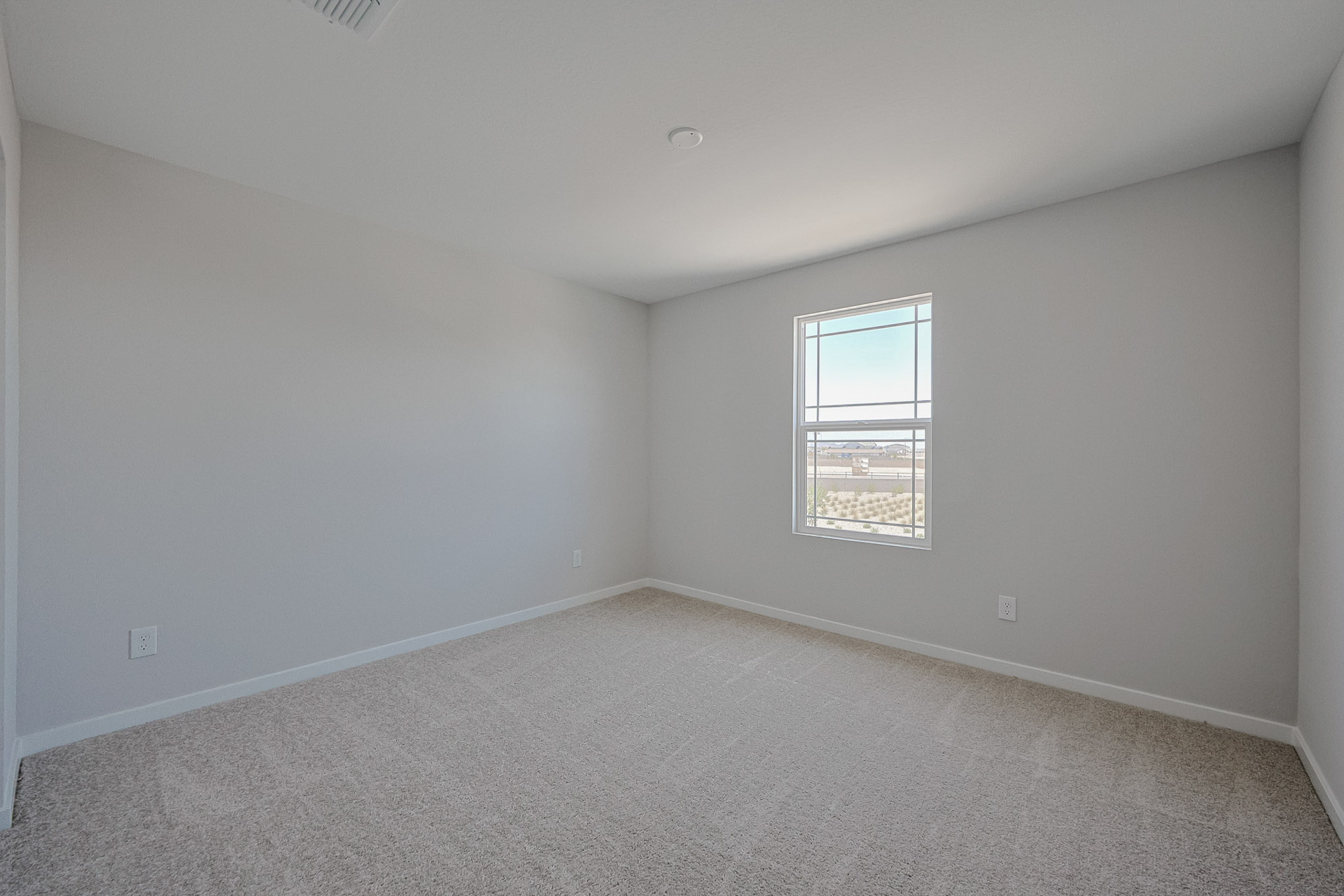 Empty room with light gray walls, beige carpet, a single window, and no furniture or decorations. Bright natural light enters through the window.