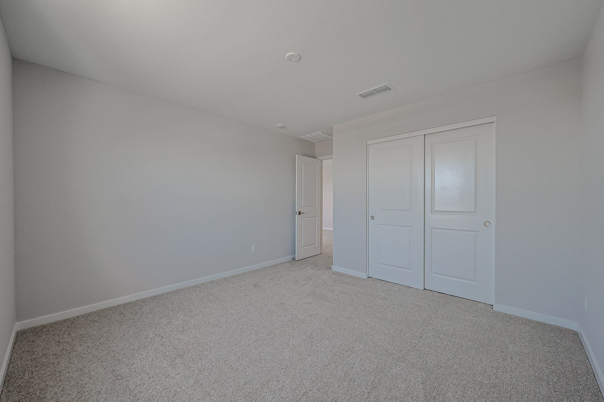 Empty room with beige carpet, light gray walls, a white door, and a double-door closet; ceiling vent and light fixture are visible.