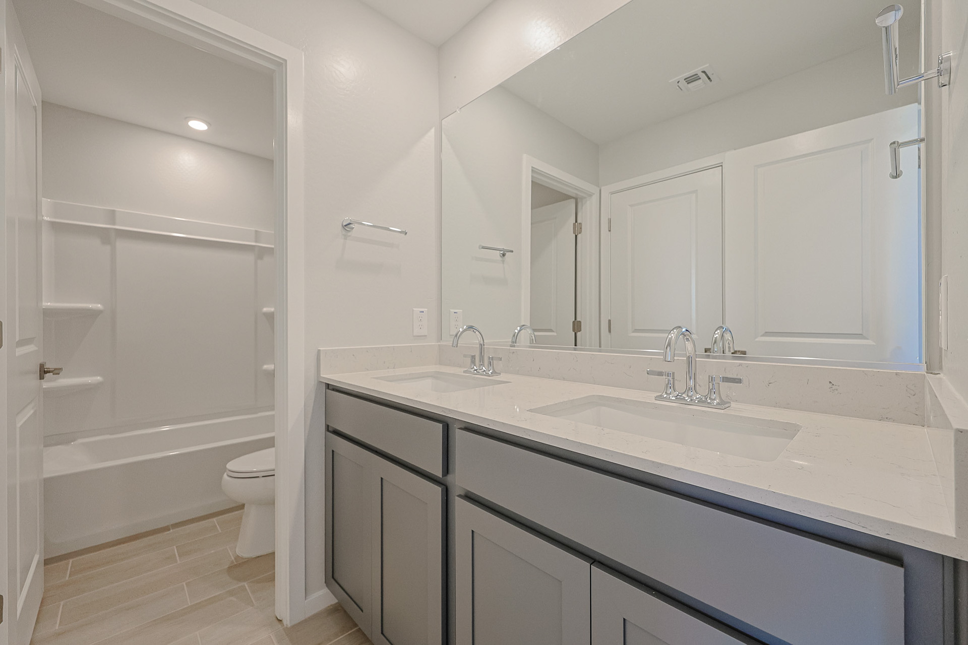 Modern bathroom with dual-sink vanity, marble countertop, large mirror, toilet, and bathtub with built-in shelves. Light gray cabinets and neutral tile flooring.