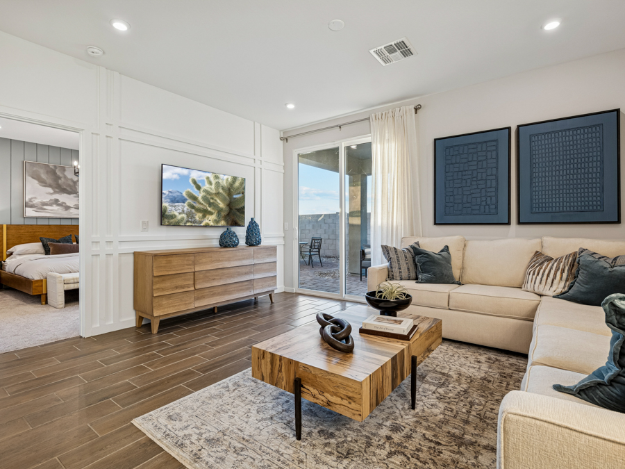 Modern living room with beige sectional sofa, wooden coffee table, TV on a wooden console, large rug, and sliding glass doors leading to a patio. Bedroom visible through open doorway.