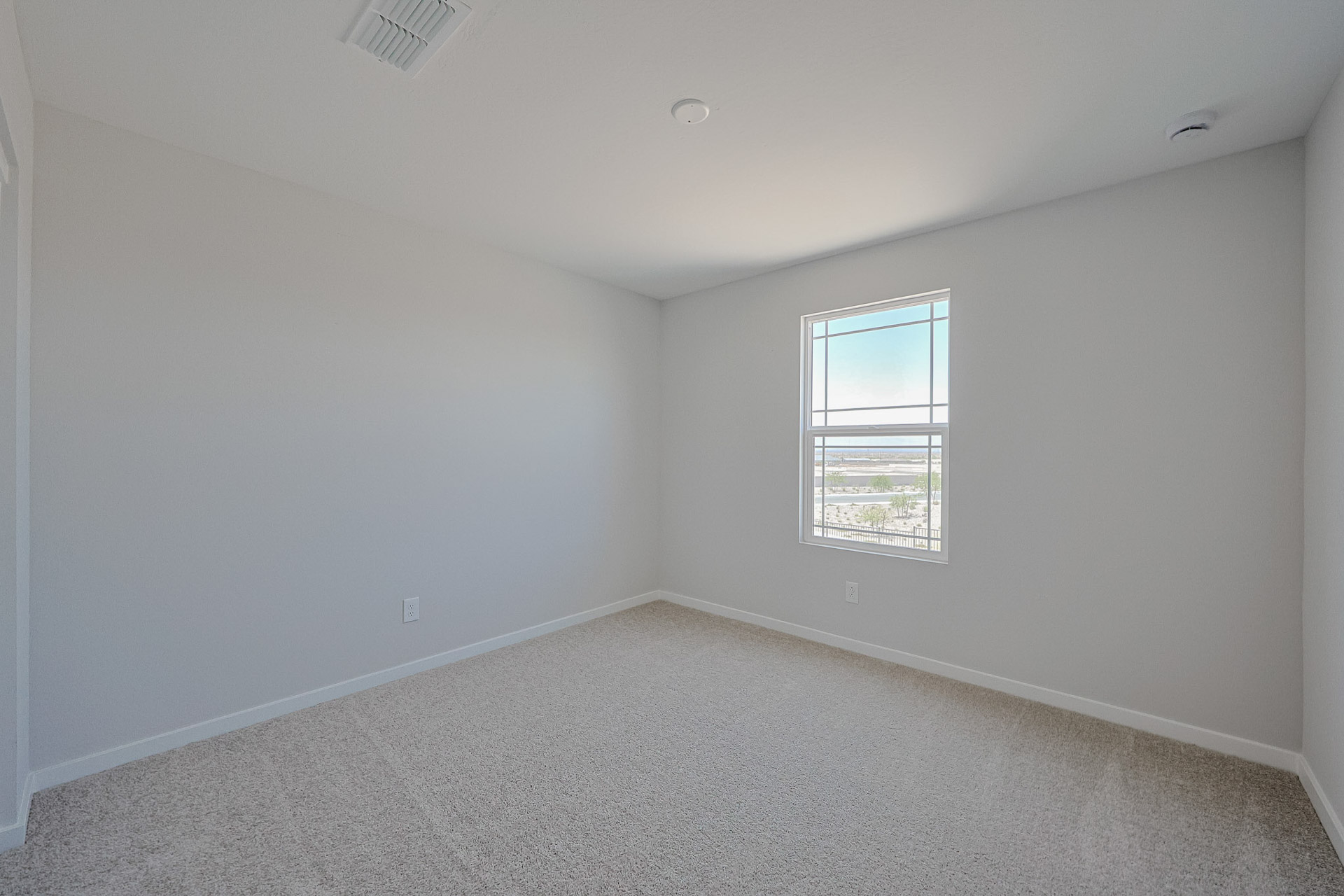 Empty room with light gray walls, beige carpet, a single window letting in natural light, and minimal fixtures visible on the ceiling and walls.