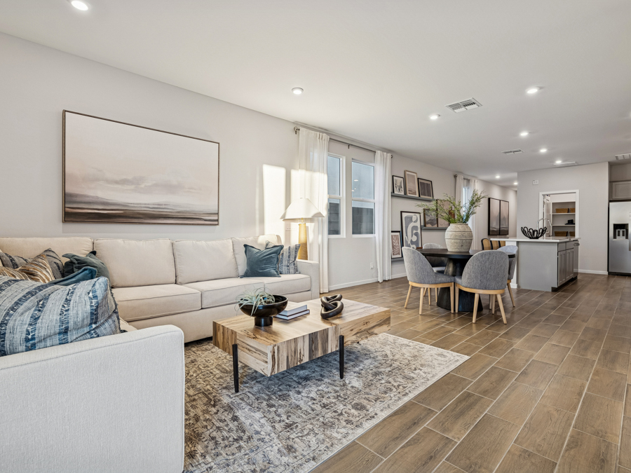 Modern open-concept living and dining area with beige sectional sofa, wooden coffee table, round dining table, and kitchen island on wood-look tile flooring. Large windows provide natural light.