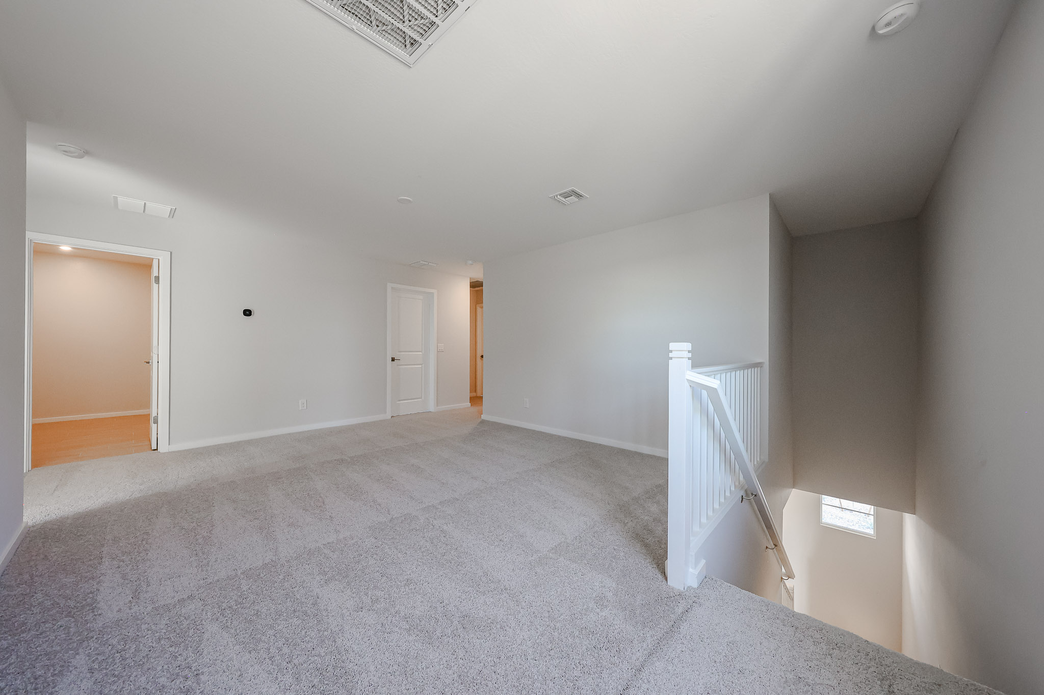 Empty carpeted room with neutral walls, white trim, and a staircase leading down; doorways visible to other rooms and natural light entering through a small window by the stairs.