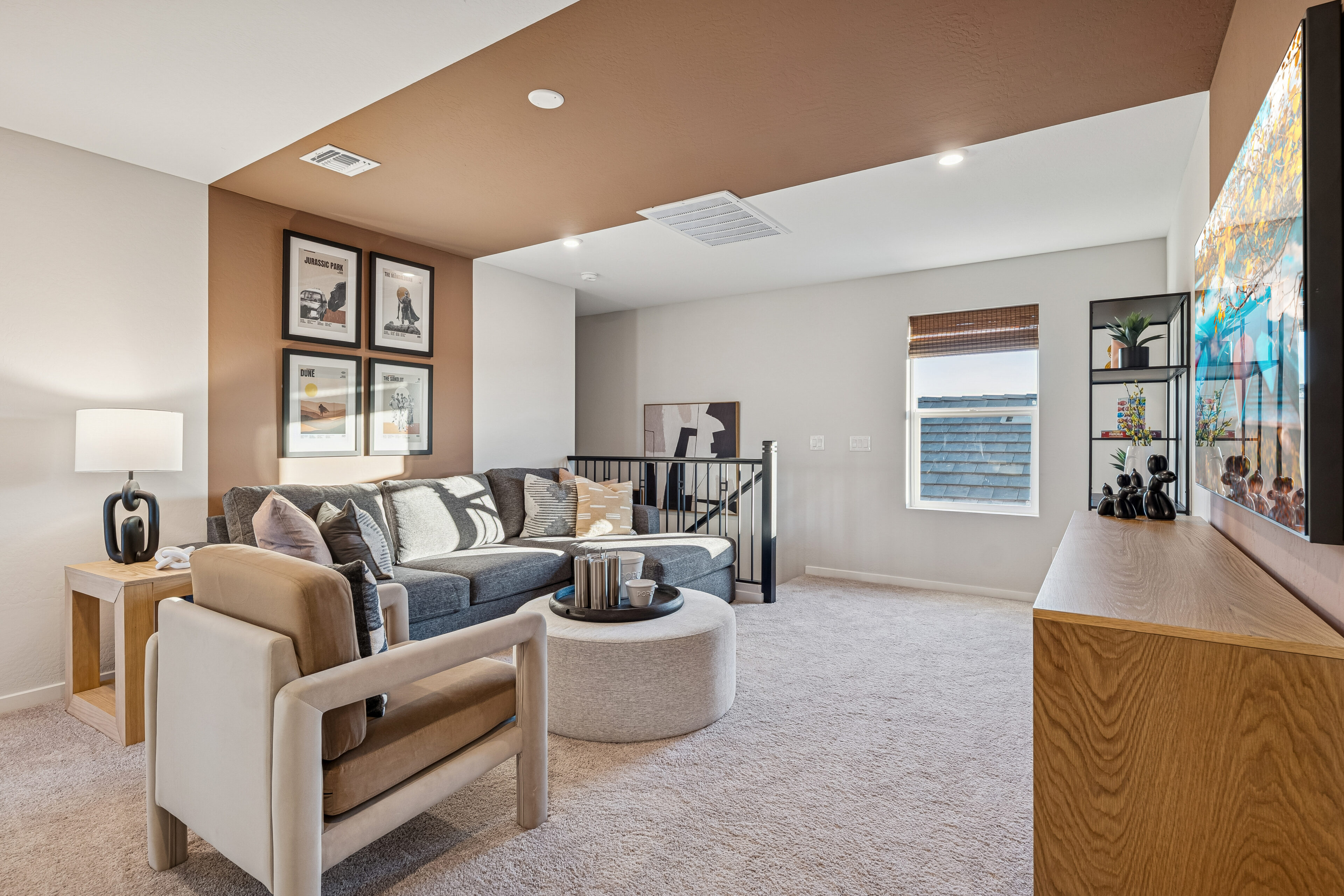 Modern living room with sectional sofa, armchair, round ottoman, wall art, TV on wooden cabinet, window with shade, and neutral decor.