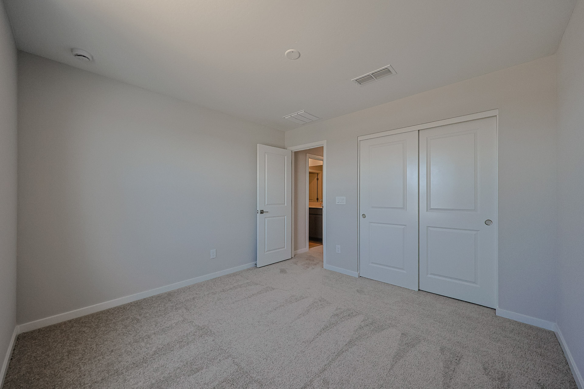 Empty bedroom with beige carpet, light gray walls, double closet doors, and an open door leading to a hallway with visible lighting.