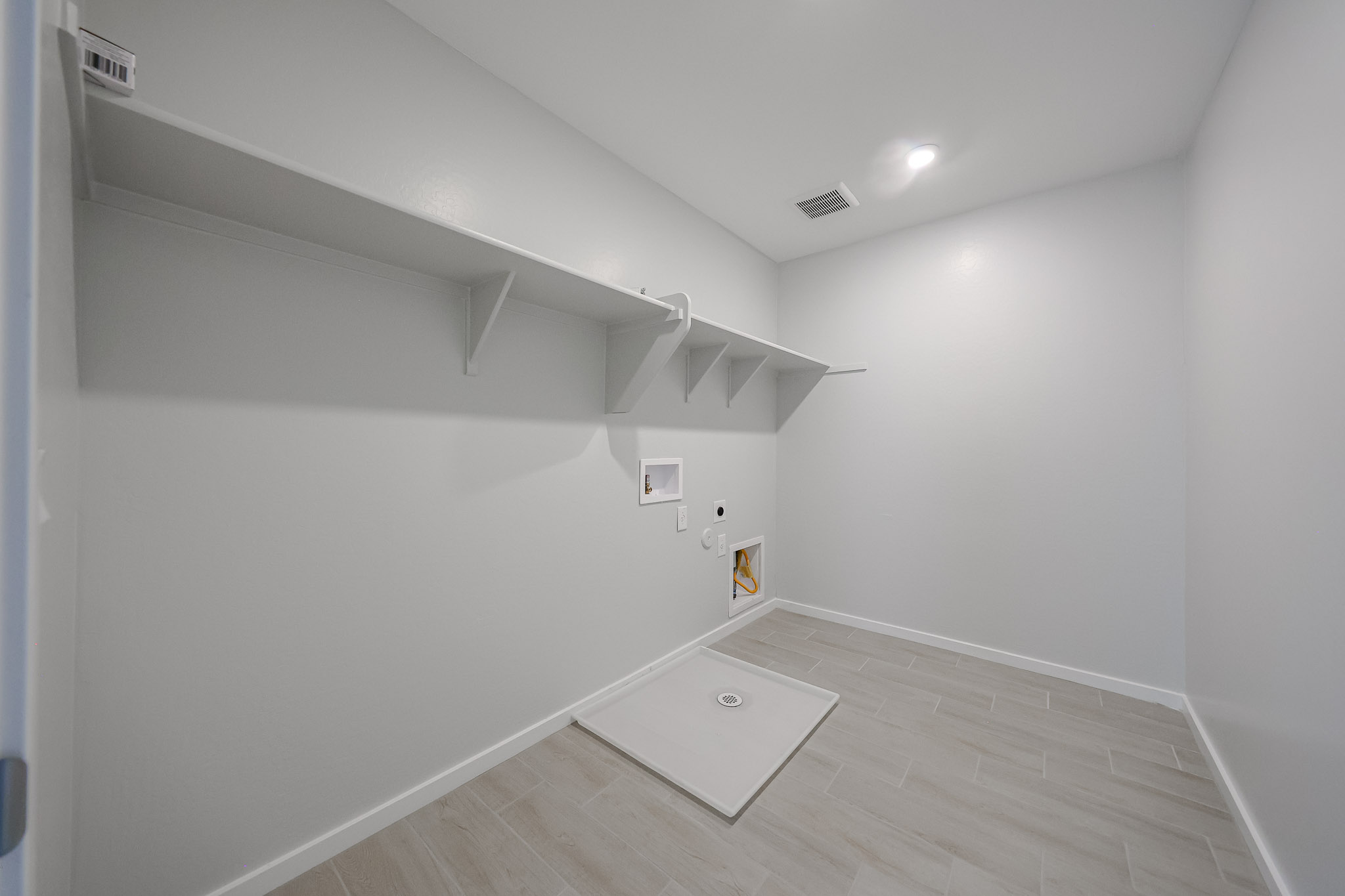 Empty laundry room with tiled floor, built-in shelves, water hookups, drain pan, and recessed lighting. Walls and ceiling are painted white.