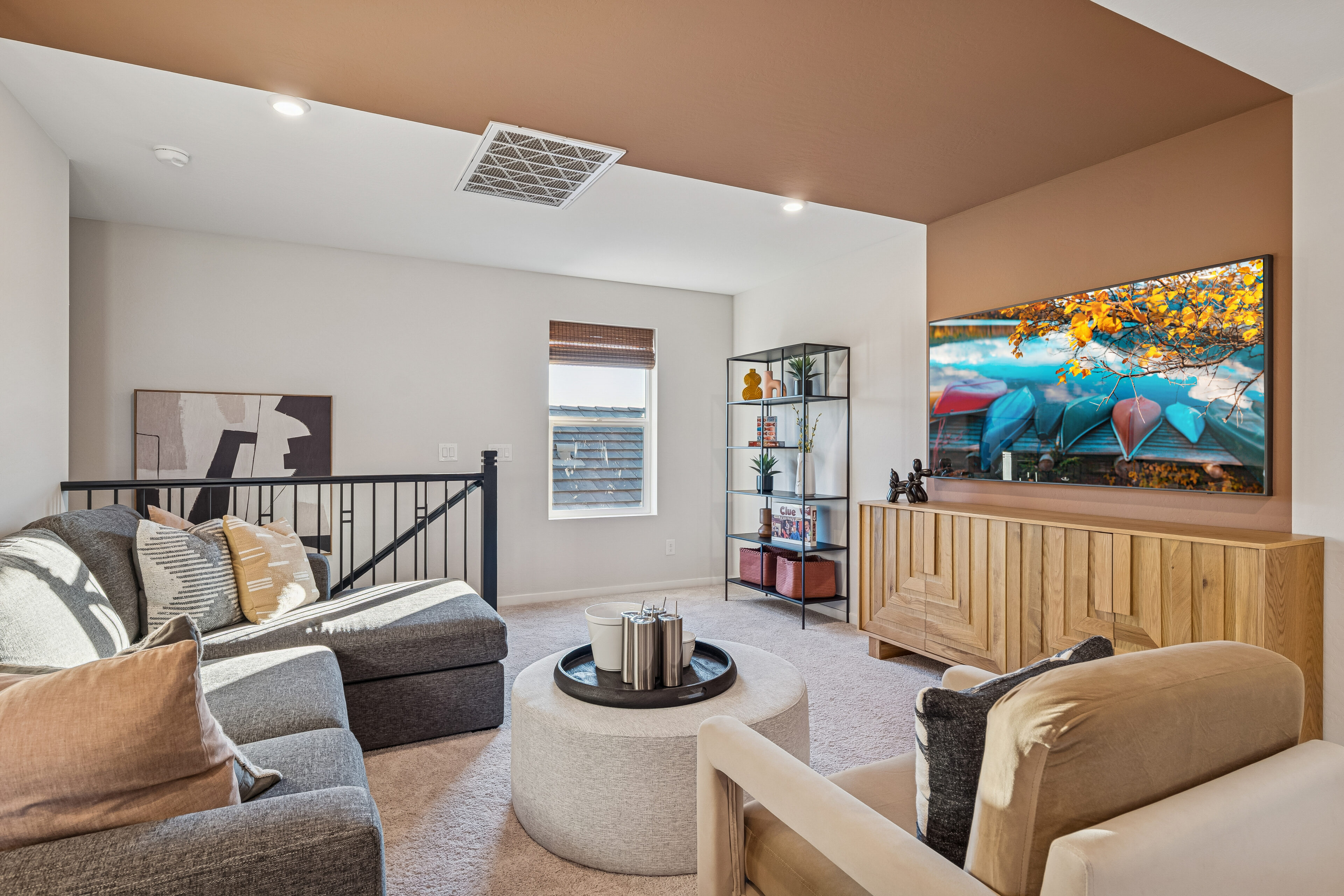 Modern living room with a sectional sofa, armchair, round ottoman, wall-mounted TV displaying autumn leaves, bookshelf, abstract art, and a window with natural light.
