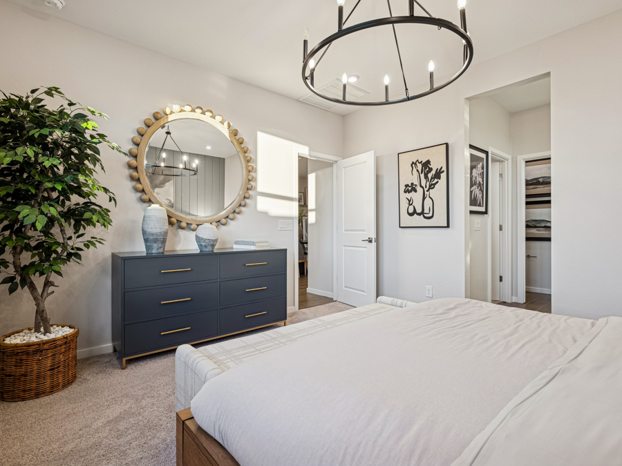 Modern bedroom with a gray dresser, round mirror, potted plant, wall art, chandelier lighting, and an open door leading to a closet.