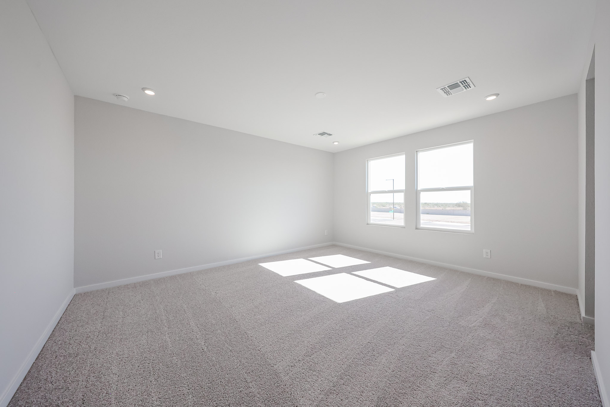 Empty, unfurnished room with light gray carpet, white walls, two large windows letting in sunlight, and ceiling lights.