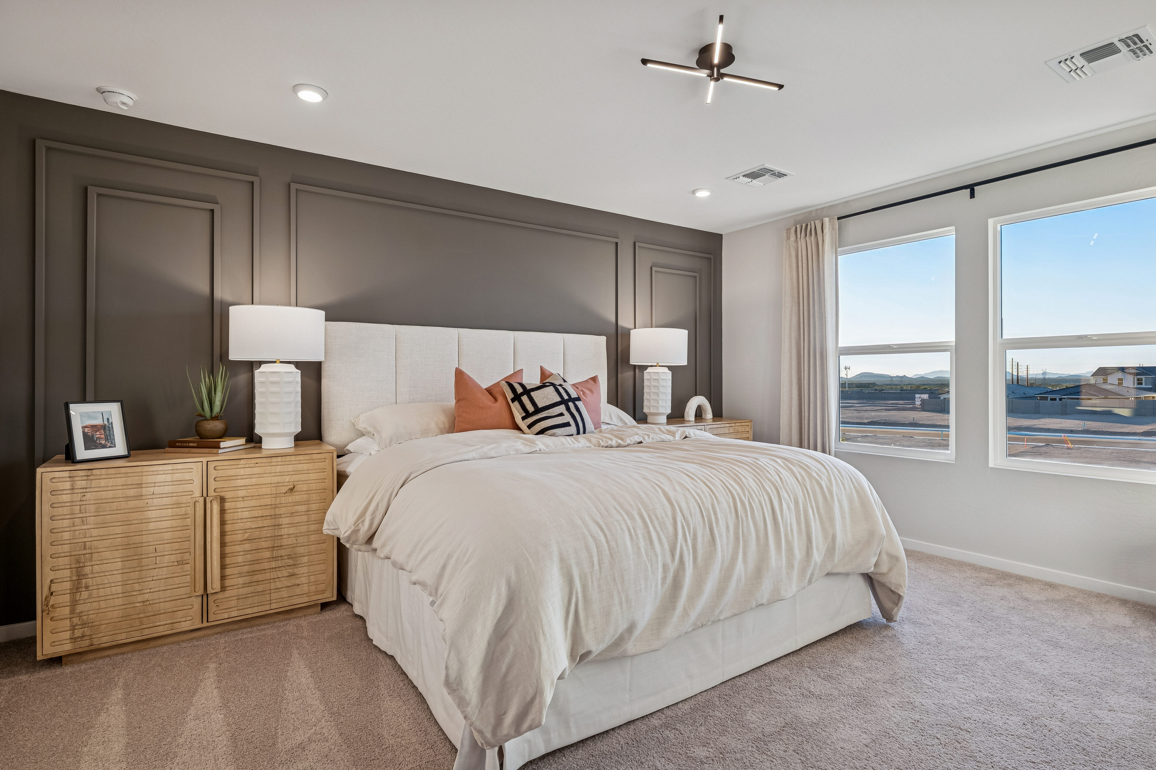 Modern bedroom with a large bed, neutral bedding, wooden nightstands, two lamps, decorative pillows, and large windows showing an outdoor view.