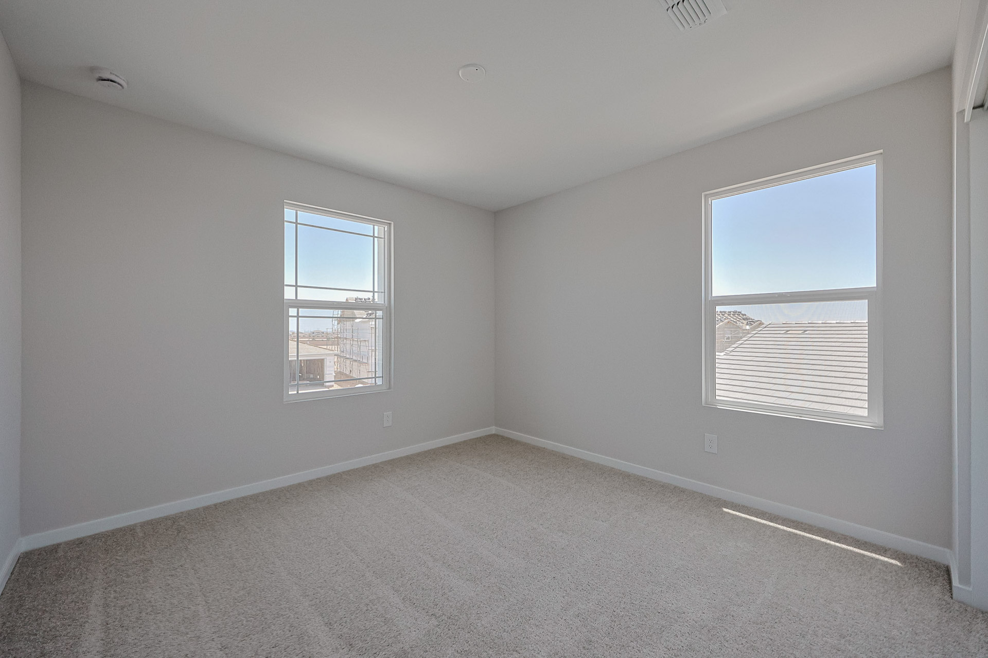 Empty, carpeted room with two windows, white walls, and a ceiling vent; daylight streams through the windows, illuminating the space.