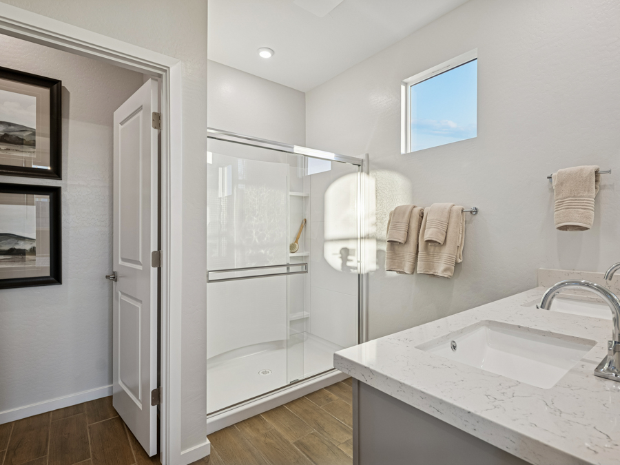 Modern bathroom with a glass shower, white countertop sink, beige towels on racks, wood-look tile floor, and two framed pictures on the wall beside an open door.