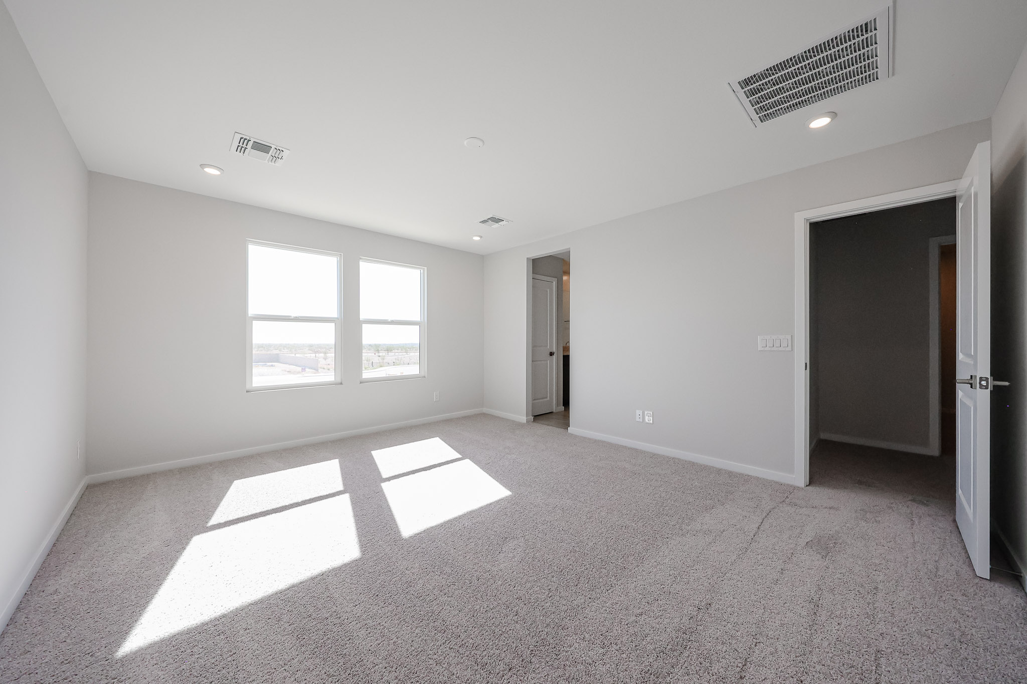 Empty carpeted bedroom with white walls, two large windows letting in natural light, and three doorways leading to other rooms. Ceiling has vents and recessed lighting.