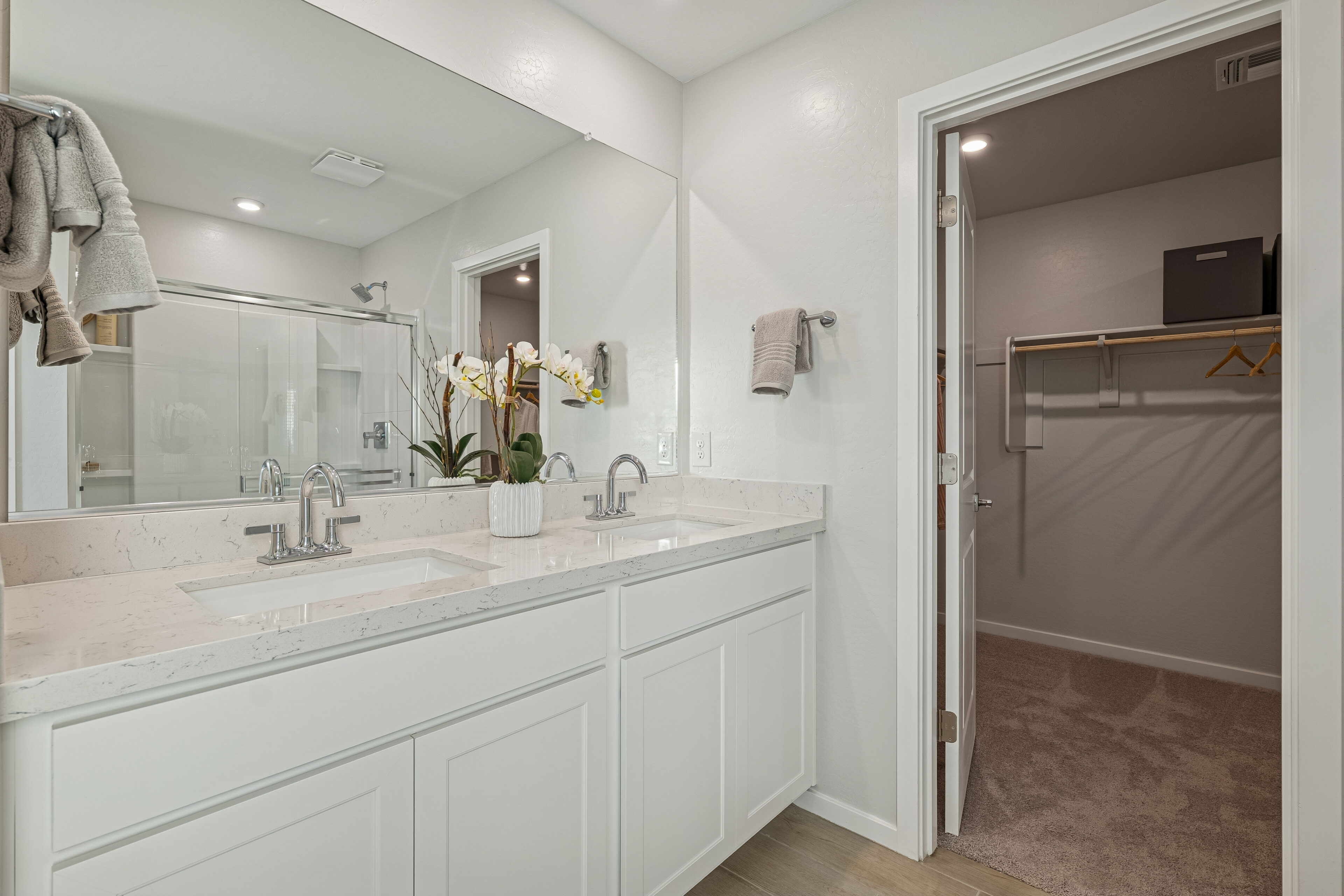 Modern bathroom with dual sinks, large mirror, white cabinets, and a potted orchid. An open door leads to a carpeted walk-in closet with shelves and hanging rods.