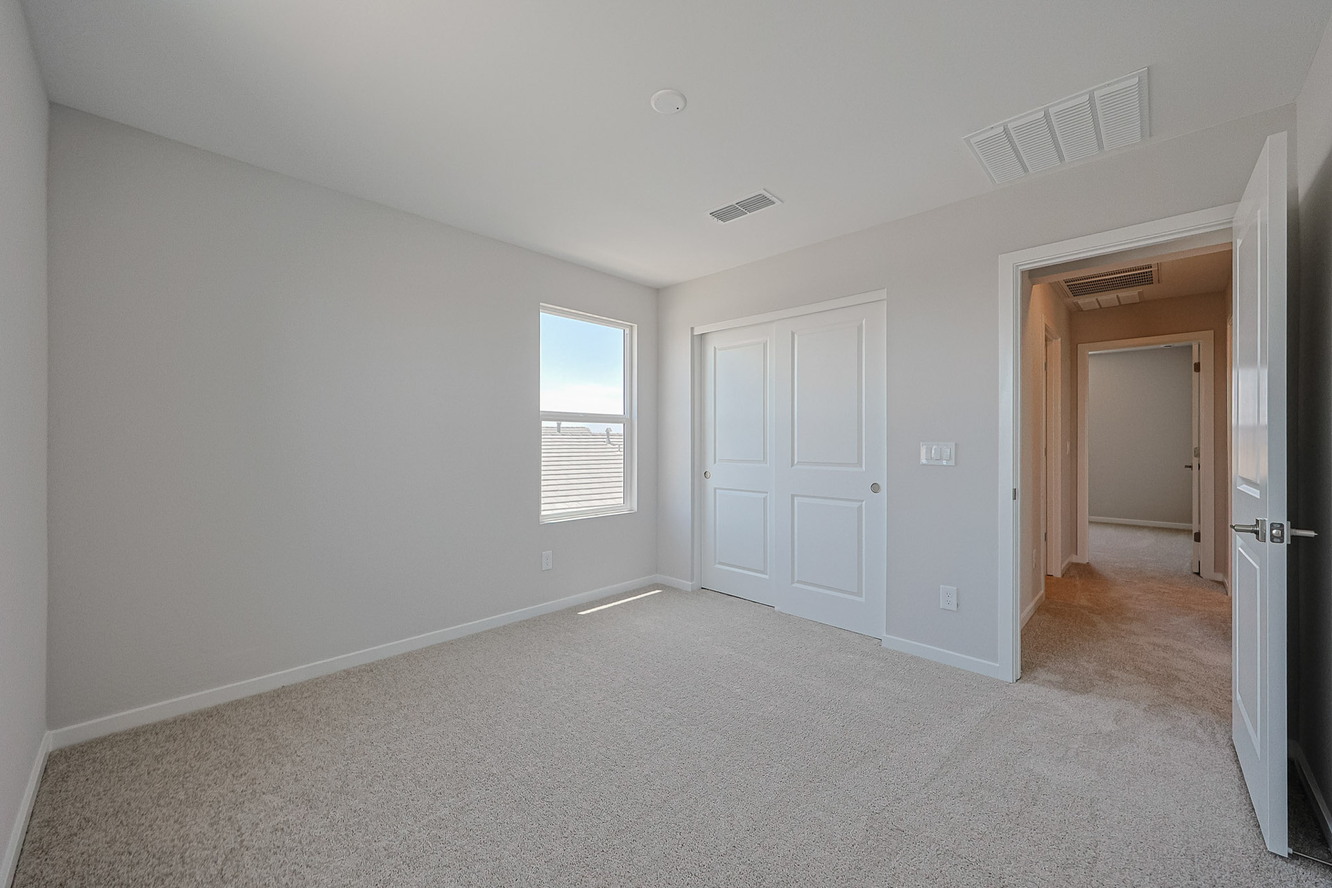 Empty bedroom with light carpet, white walls, a window, double closet doors, and an open door leading to a hallway.