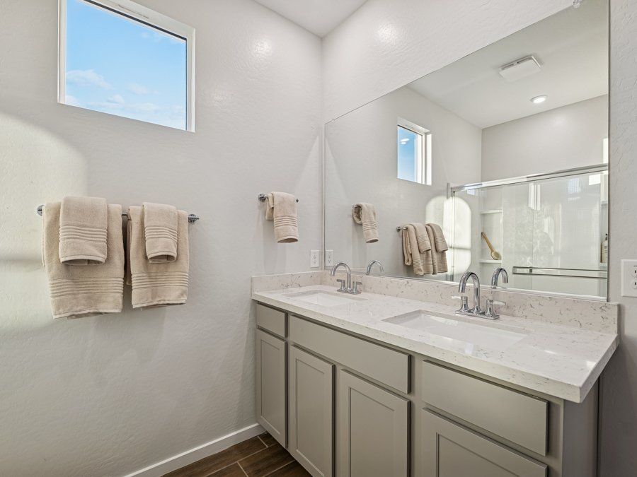 Modern bathroom with double sinks, large mirror, gray cabinets, marble countertop, beige towels, and walk-in shower with glass doors. Natural light from two small windows.