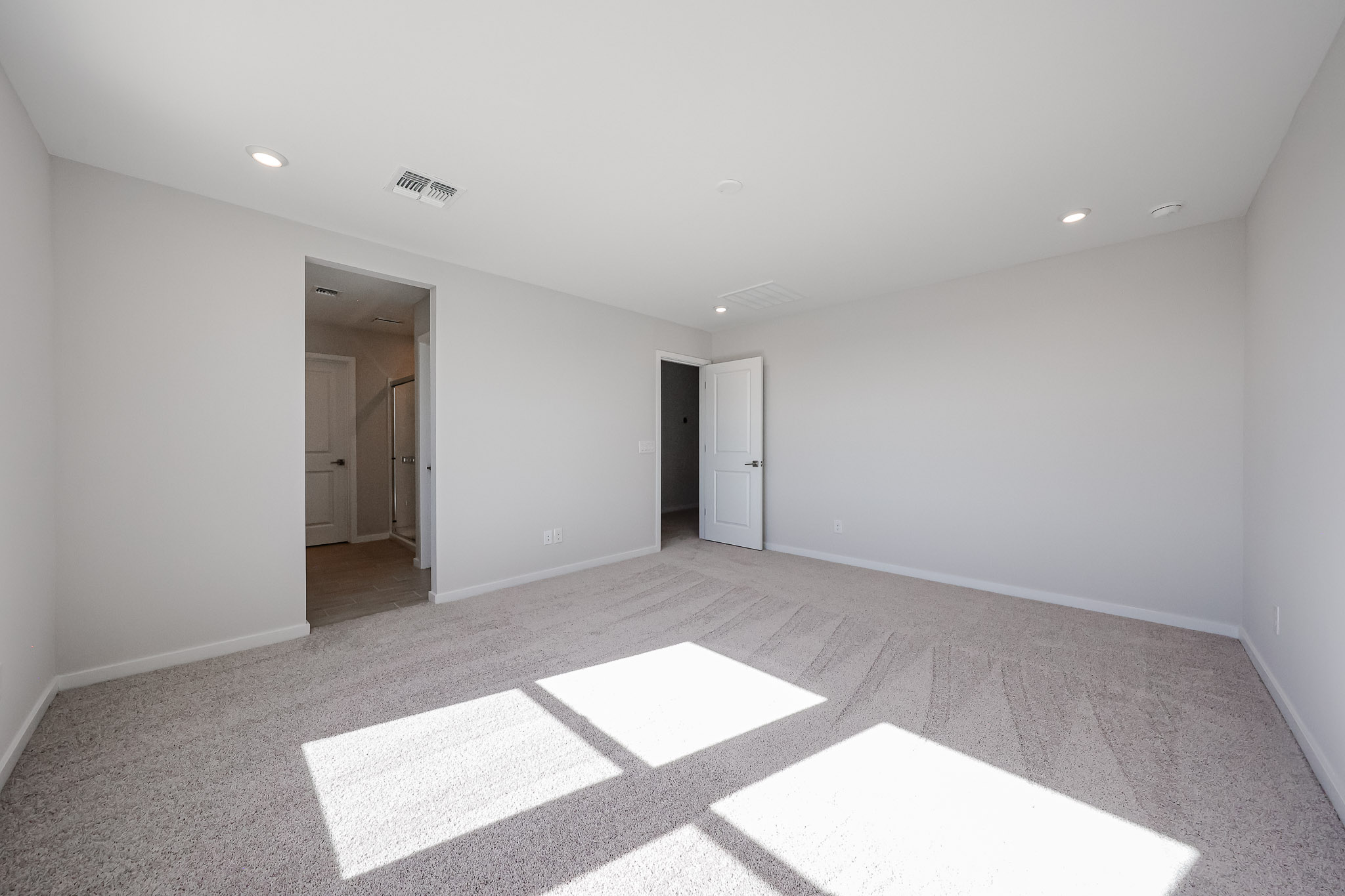 Empty, carpeted bedroom with white walls, recessed ceiling lights, two open doors, and sunlight streaming through a window onto the floor.