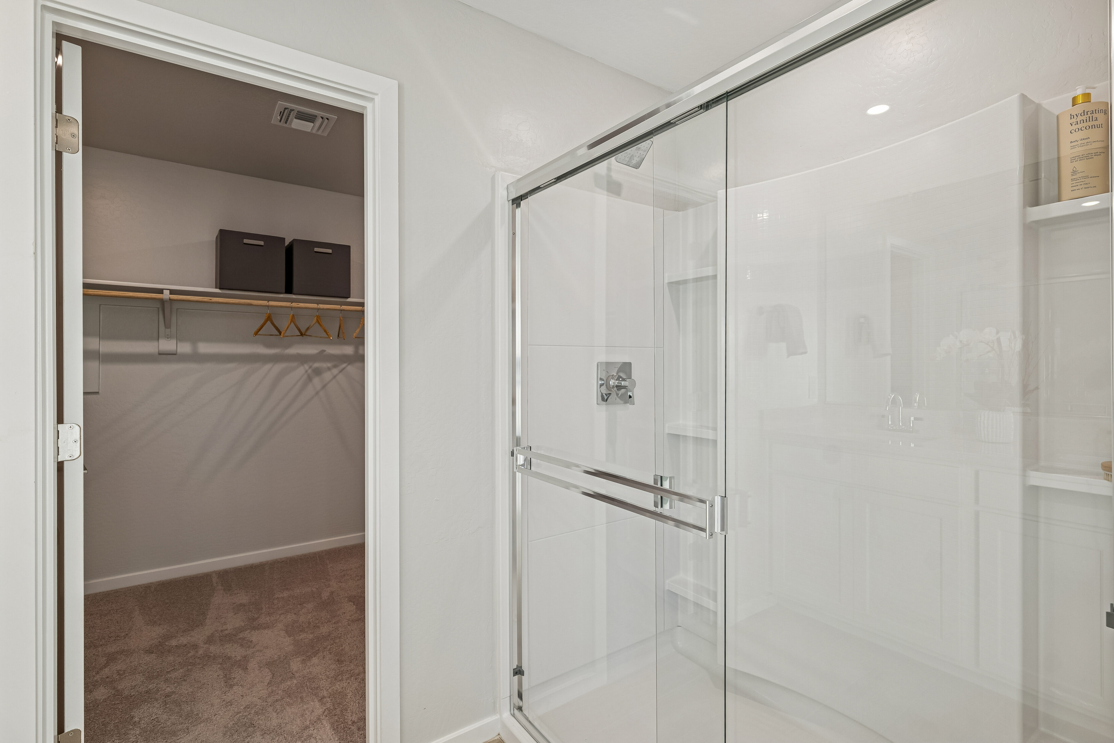Walk-in closet with shelves and hangers next to a bathroom with a glass shower enclosure and a white vanity.