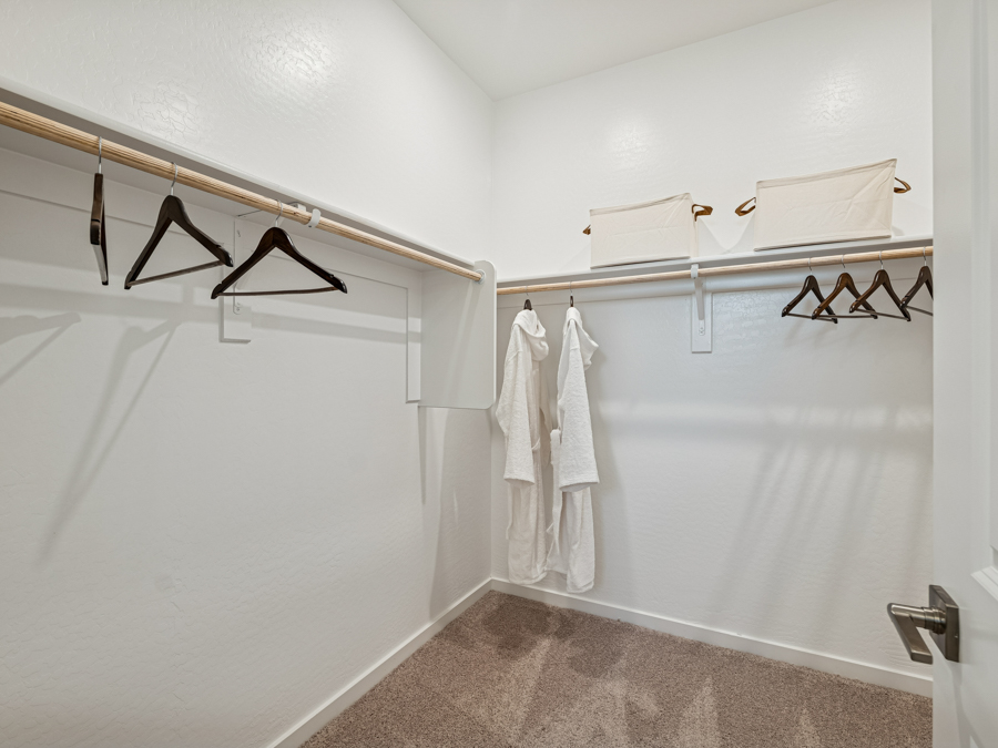 Empty walk-in closet with beige carpet, white walls, wooden hanging rods, a few black hangers, two white robes, and two white storage bins on the upper shelf.