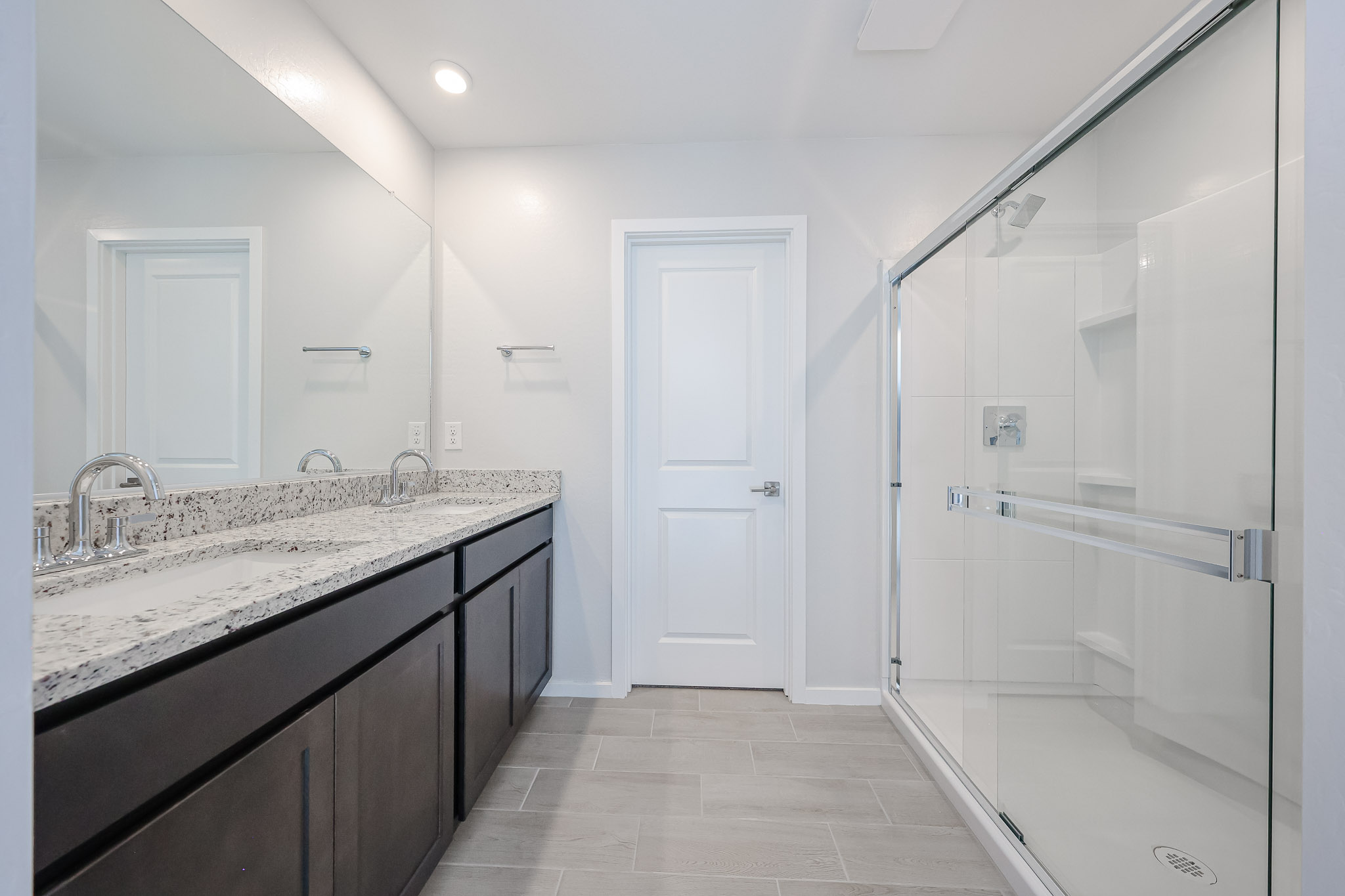 Modern bathroom with a double-sink vanity, granite countertop, large mirror, glass shower enclosure, and light gray tile floor.