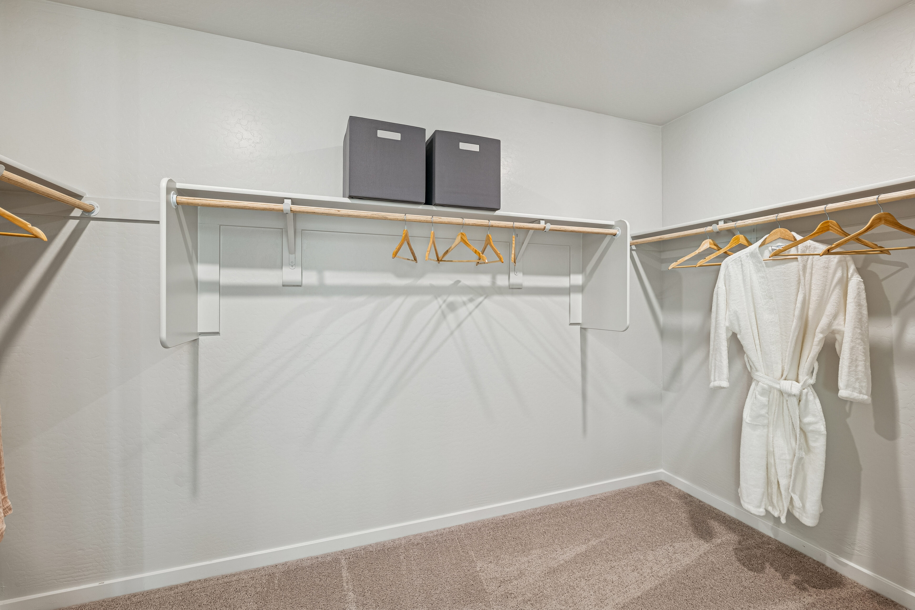Spacious walk-in closet with beige carpet, two storage boxes, several wooden hangers, and a white robe hanging on the right side.