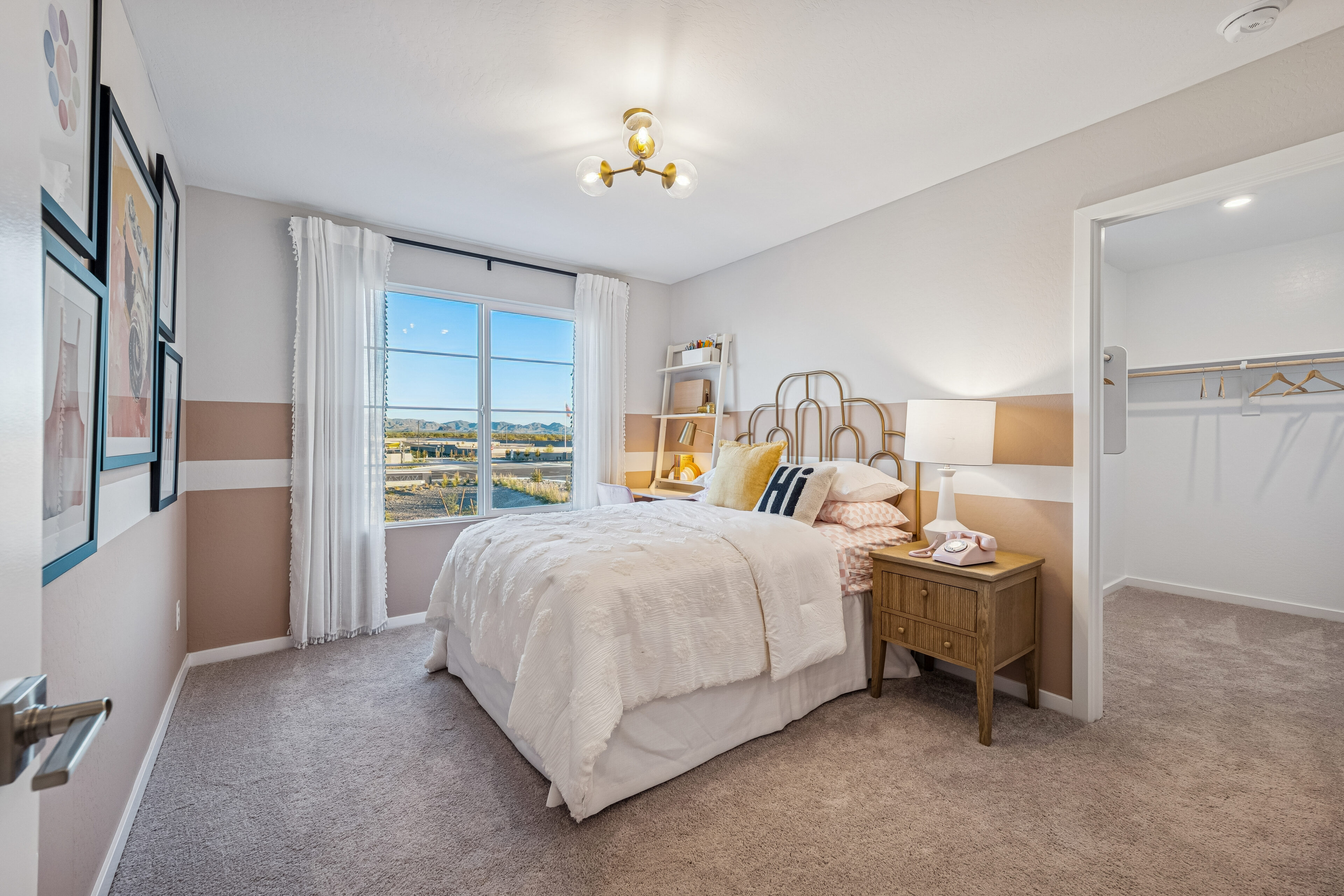 A neatly made bed with white bedding is next to a nightstand in a bright bedroom with a window, wall art, and an open walk-in closet with shelves and hangers.