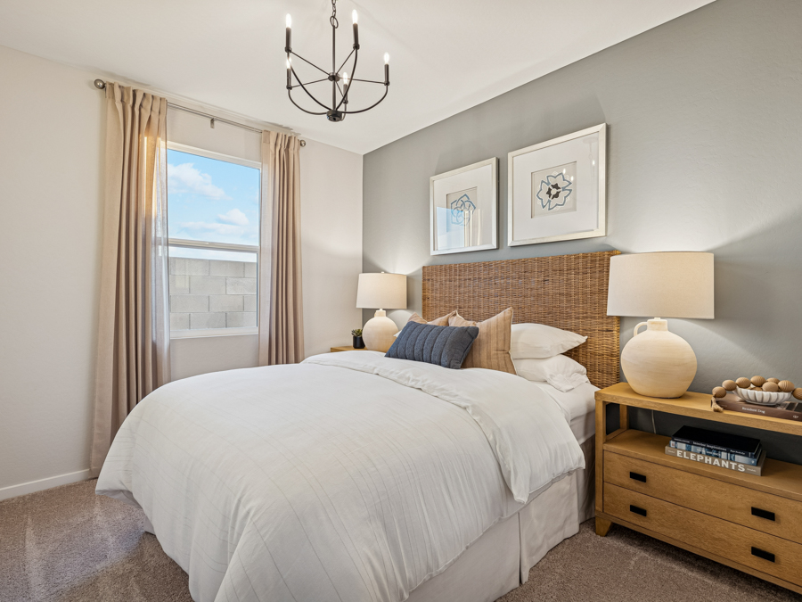Modern bedroom with a neatly made bed, two wooden nightstands with lamps, framed art above the headboard, and a window with beige curtains letting in daylight.