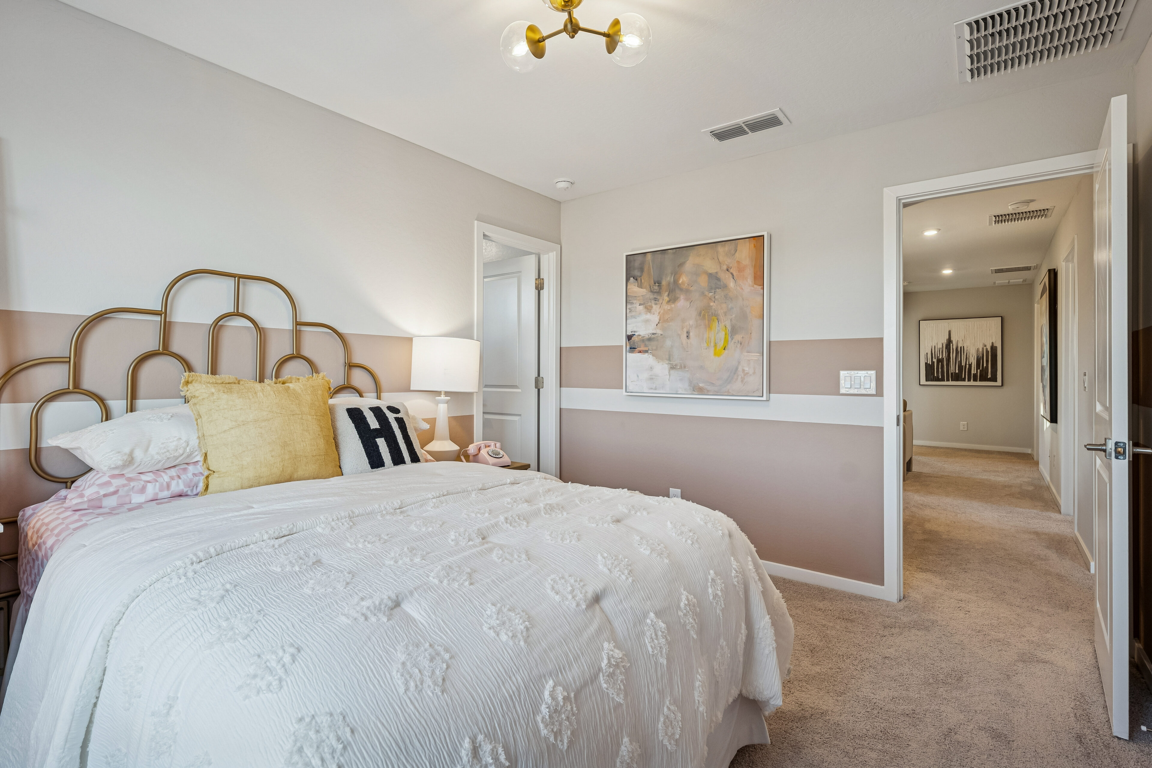 Bedroom with a white bed, decorative pillows, wall art, nightstand lamp, and neutral-toned walls. An open door reveals a hallway and additional room in the background.