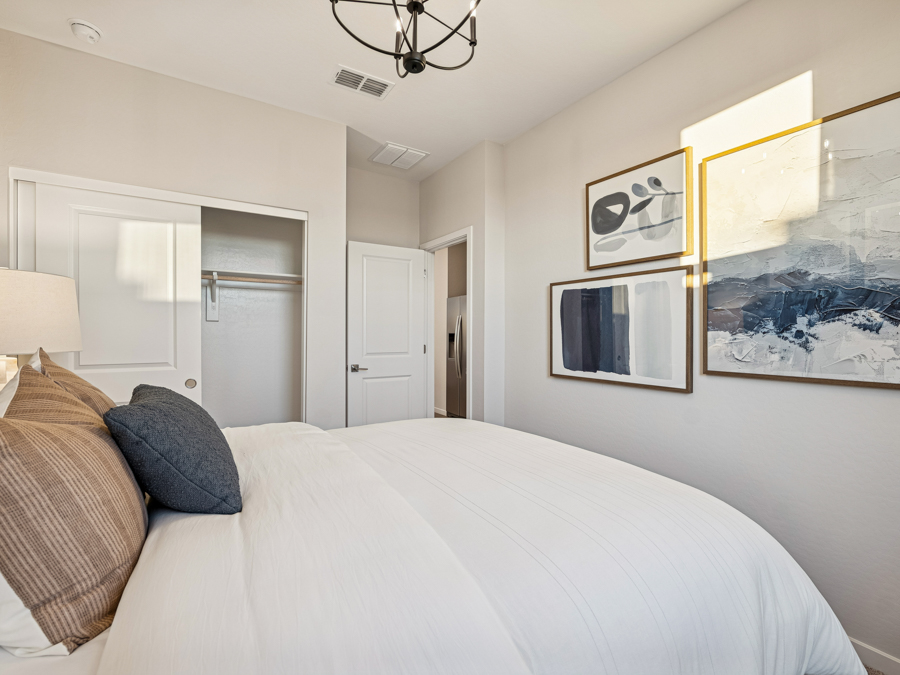 Modern bedroom with a neatly made bed, neutral bedding, wall art, an open closet, and white walls. Natural light enters through a window.
