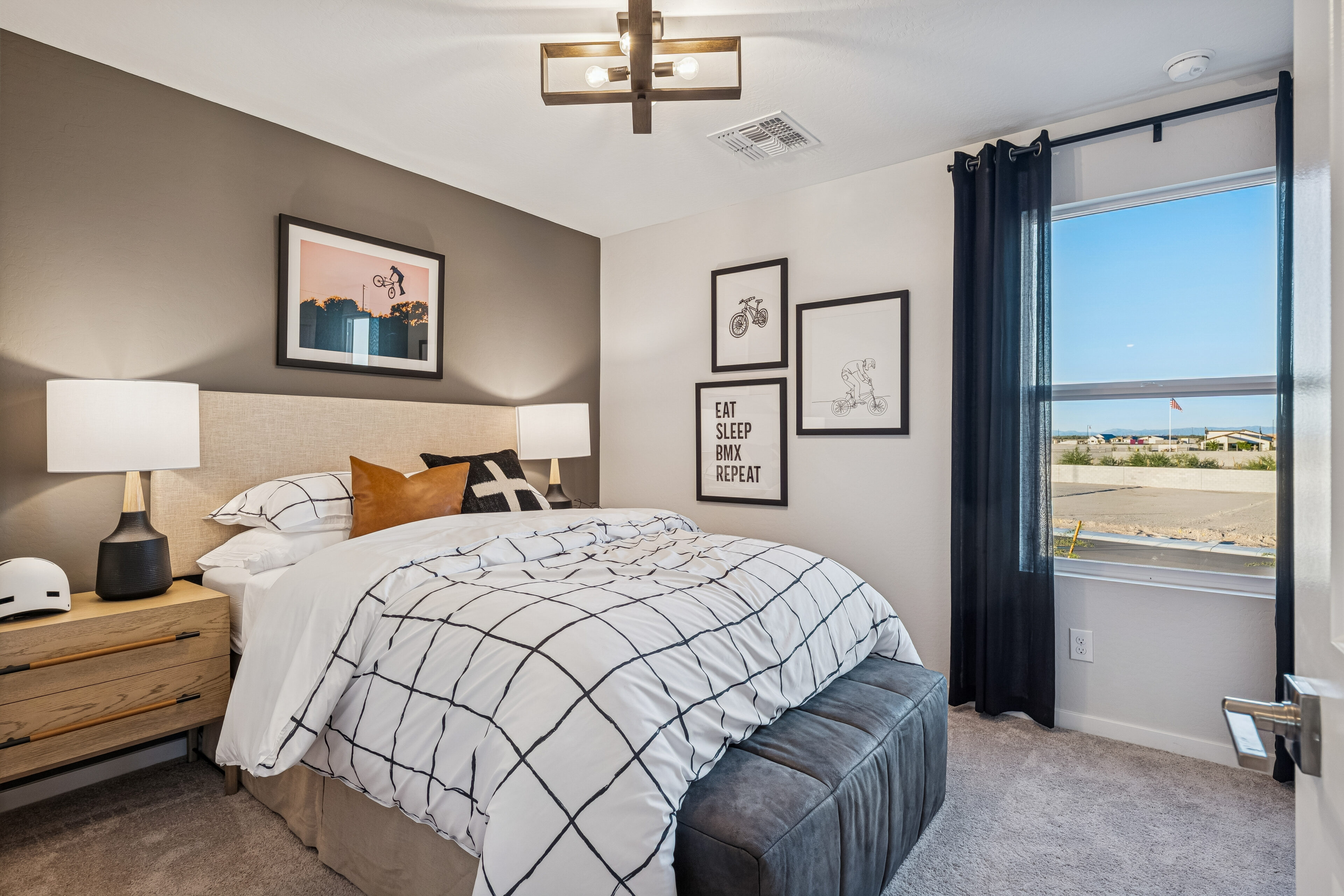 A neatly made bed with checkered bedding is in a modern bedroom, decorated with framed art, two lamps, a window with black curtains, and a view of a sunny outdoor scene.