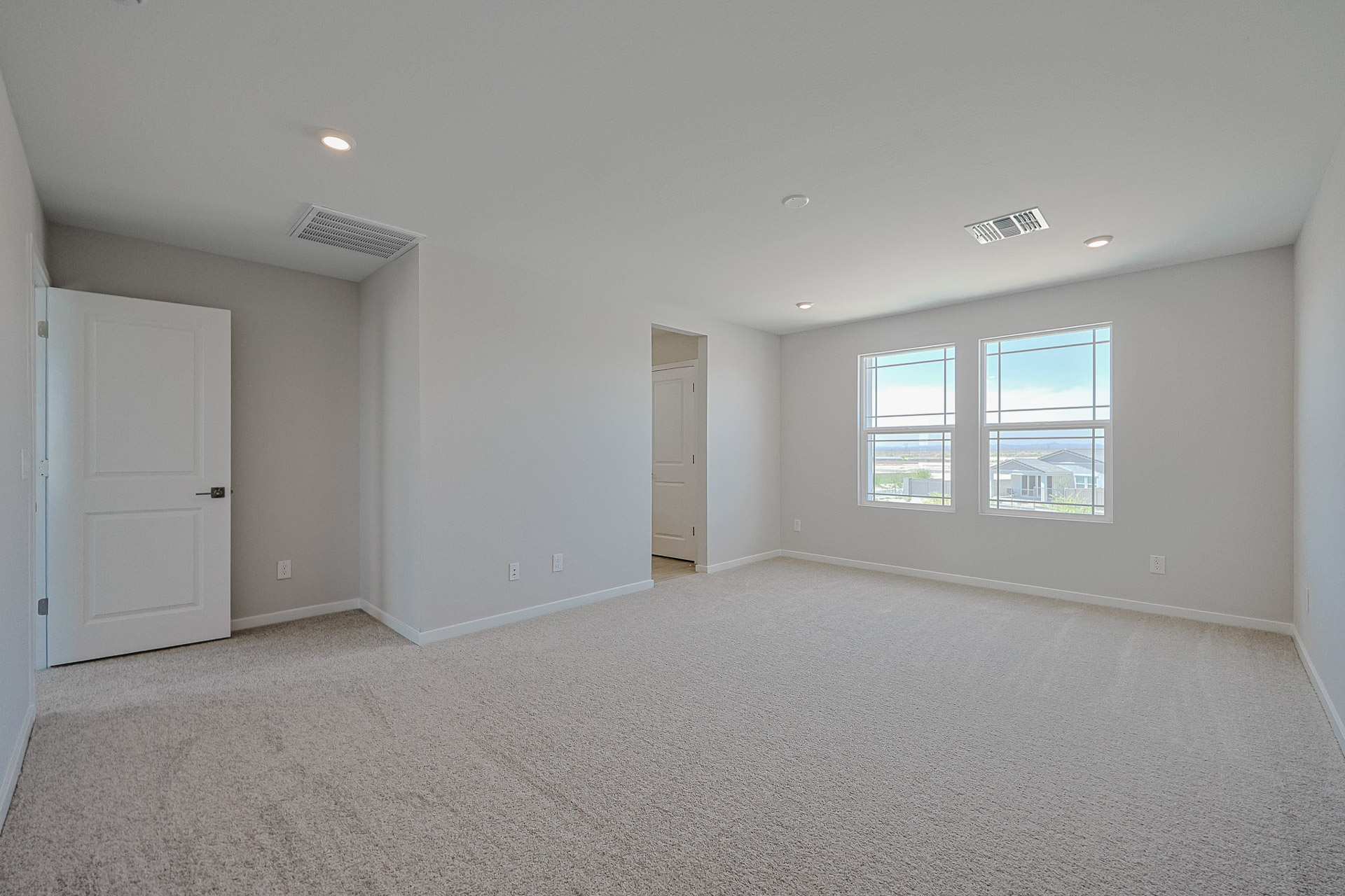 Empty, carpeted bedroom with two windows, white walls, ceiling lights, an open door, and a small open doorway leading to another area.