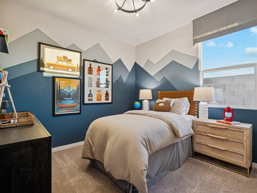 A neatly made bed in a child’s room with mountain-themed wall art, blue and white walls, and a window letting in daylight. A nightstand holds two lamps and a red lantern.