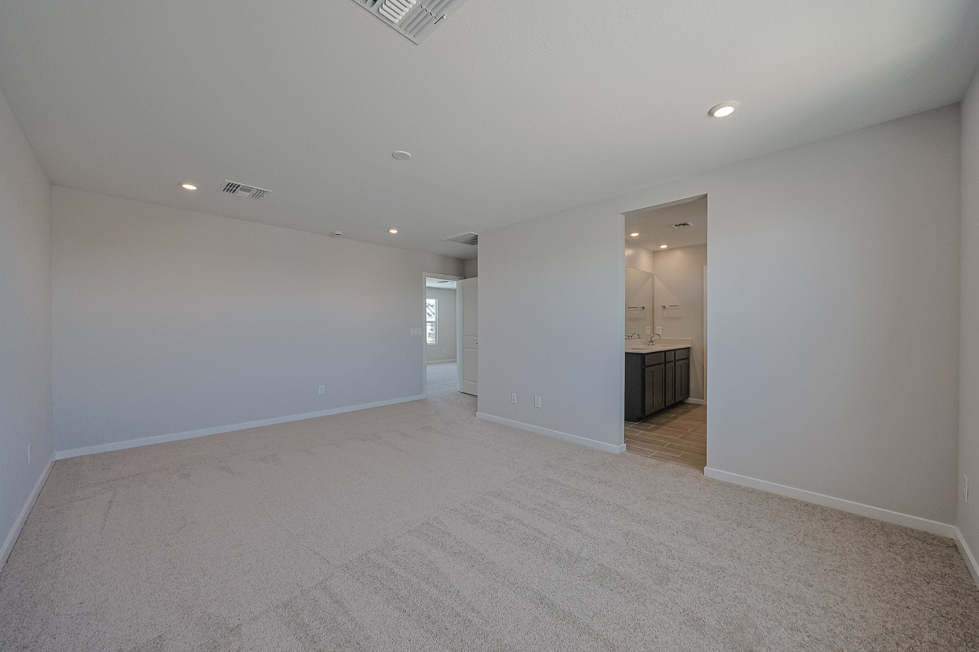Spacious, empty carpeted room with light-colored walls, ceiling lights, and an open doorway leading to a bathroom with dual sinks and large mirror. Natural light enters from a window.