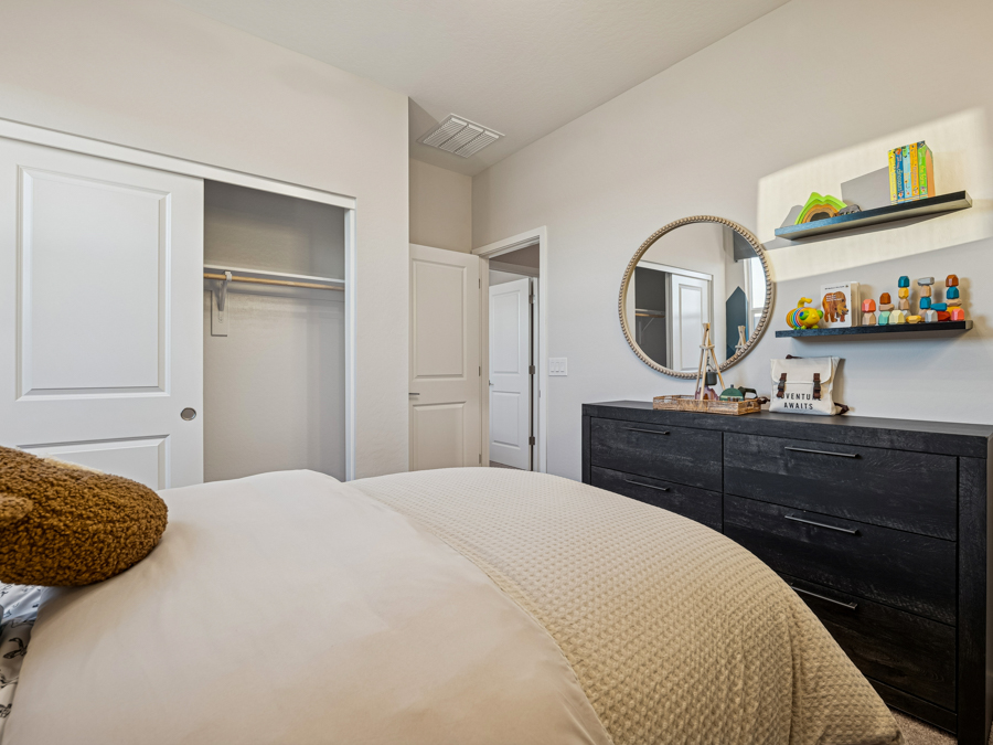 A neatly made bed faces a dark dresser with toys and shelves. An open closet with sliding doors is visible. Walls are light-colored.