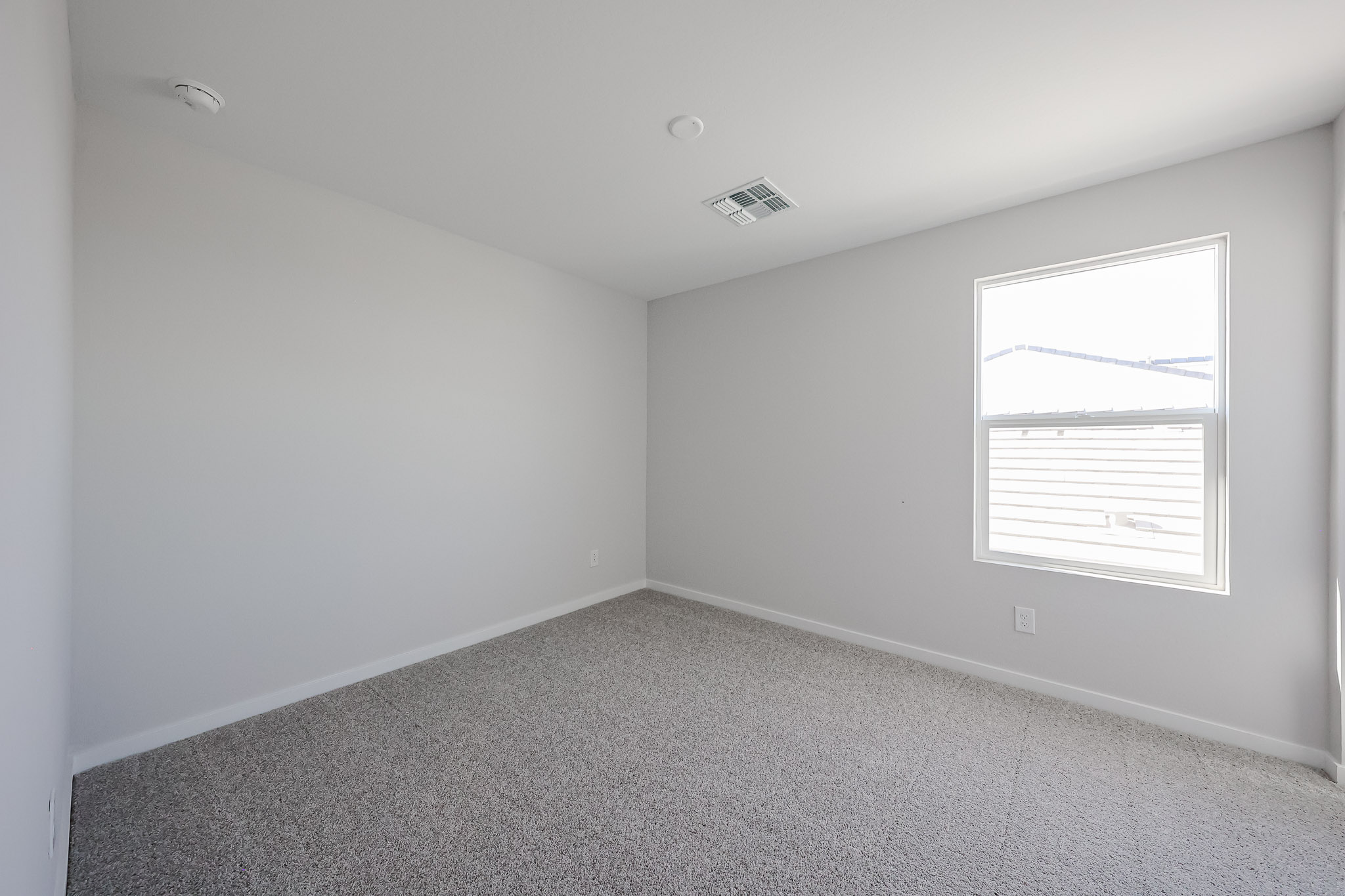 Empty room with light gray walls, carpeted floor, a single window on the right wall, and a ceiling vent.