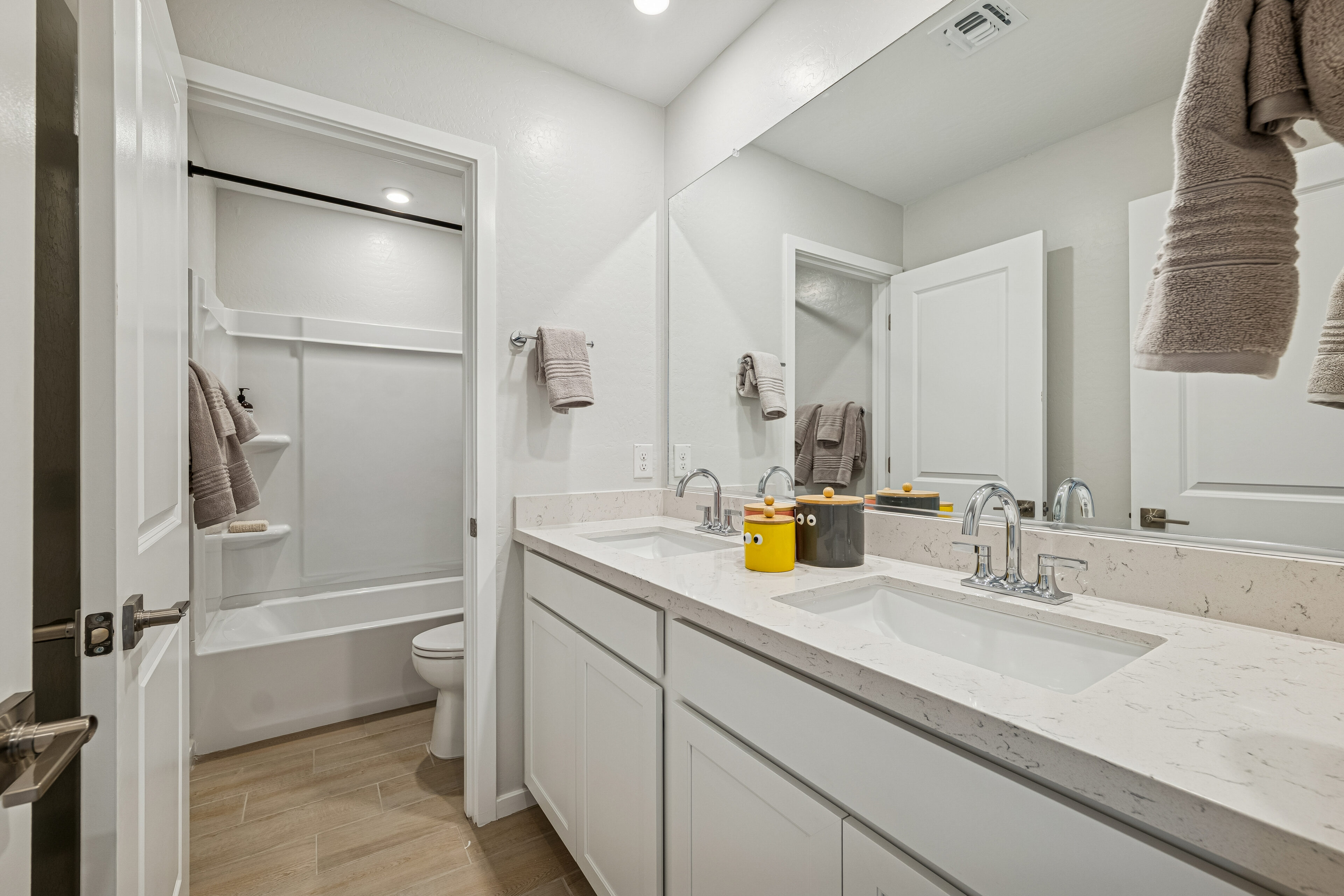 Modern bathroom with double sinks, large mirror, white cabinets, towel racks, and a bathtub with a shower curtain at the back. Neutral color scheme with beige and white tones.