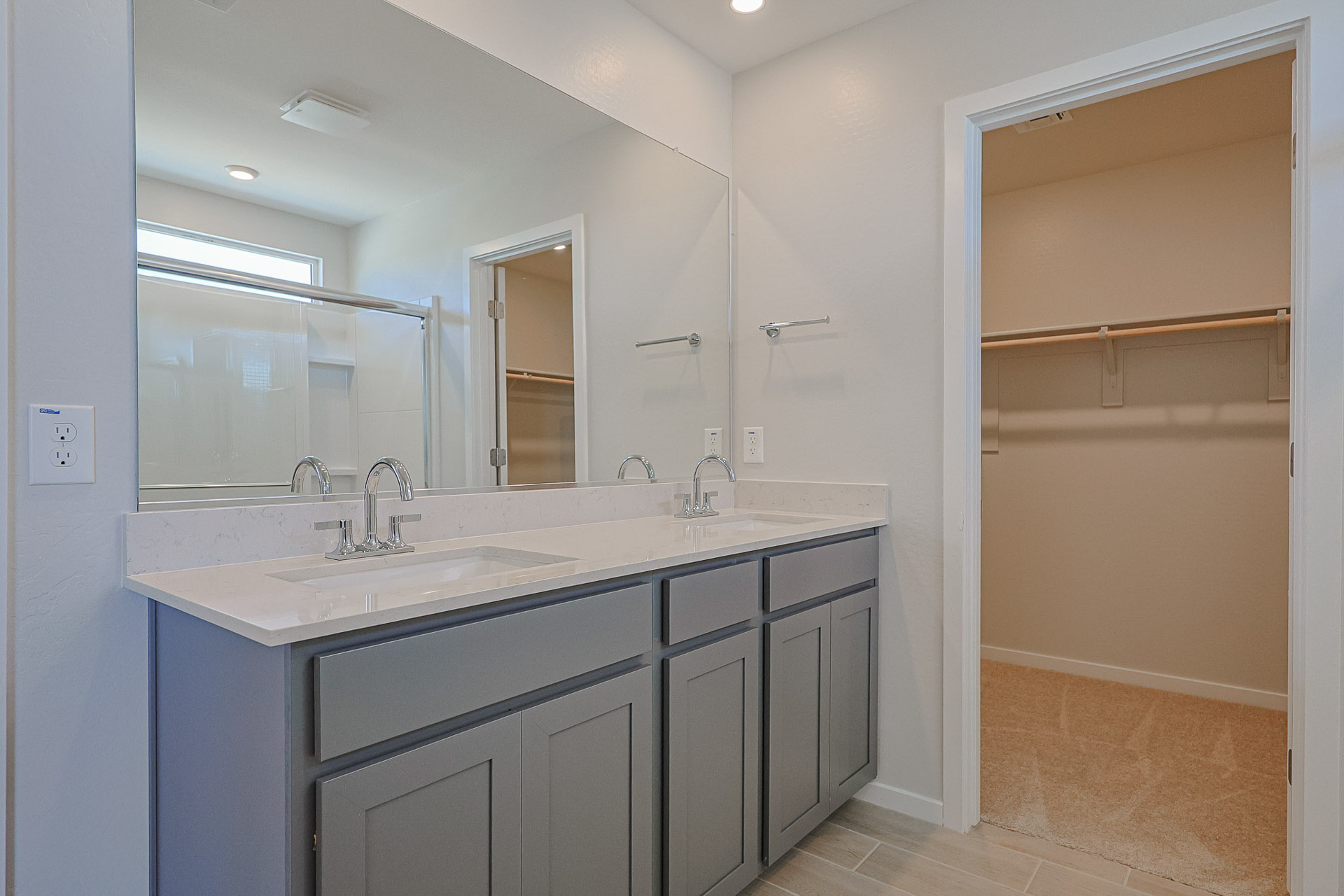 Bathroom with double sink vanity, large mirror, gray cabinets, and view into a walk-in closet with beige carpet and wooden shelves.