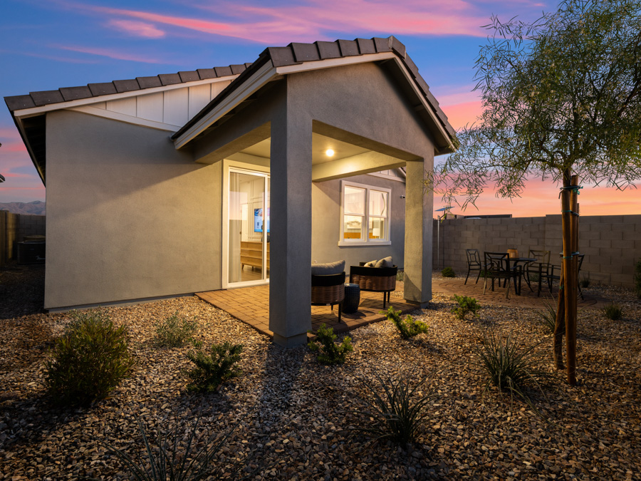 Small modern house with a covered patio, outdoor seating, and a dining table in a landscaped backyard at sunset.