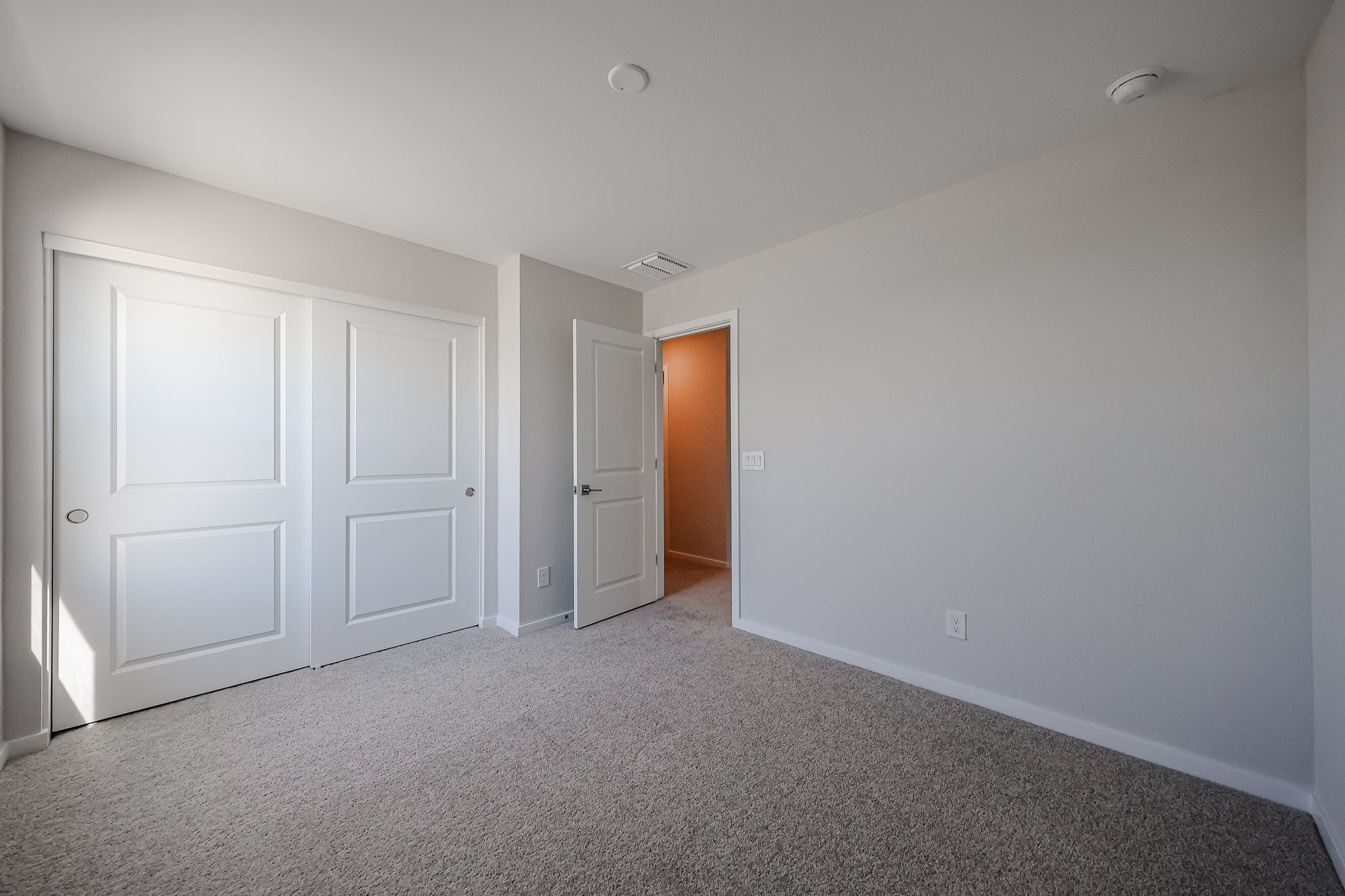 Empty bedroom with light gray walls, beige carpet, a double-door closet, and an open door leading to a hallway.