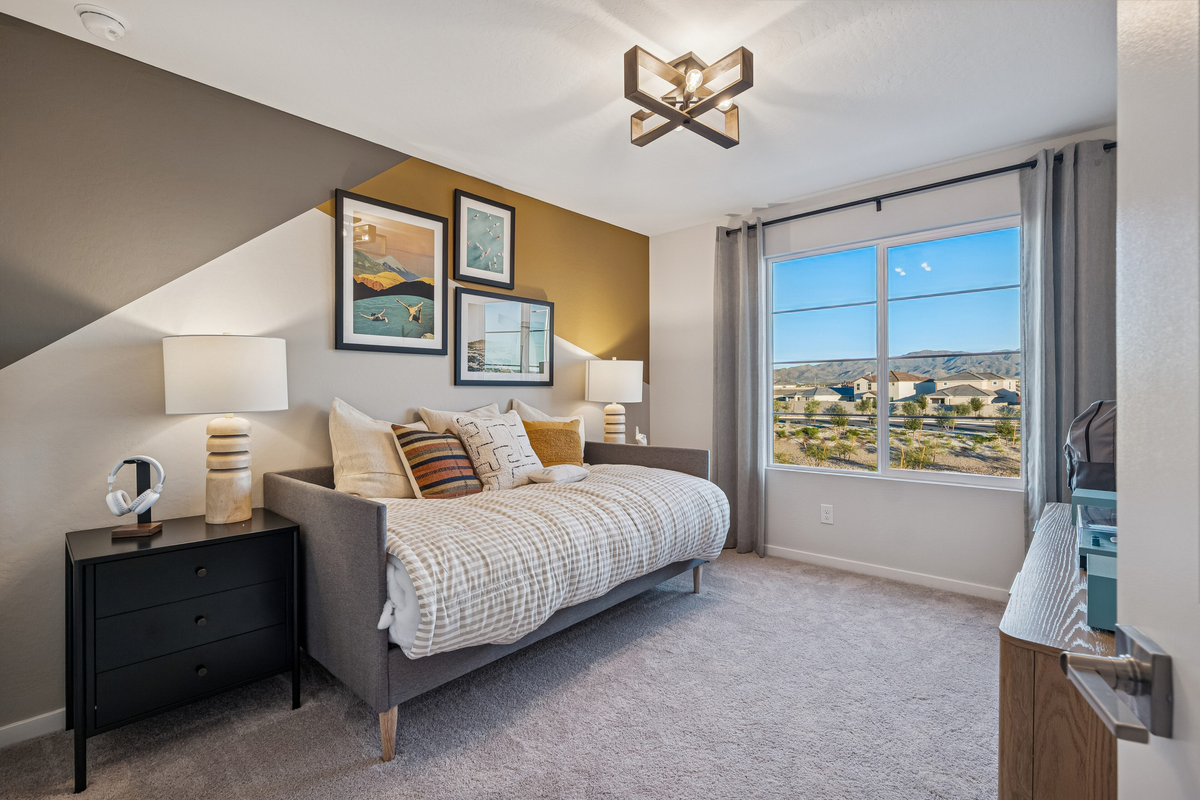 Modern bedroom with a geometric accent wall, daybed, two lamps, wall art, and a large window overlooking scenic houses and mountains.