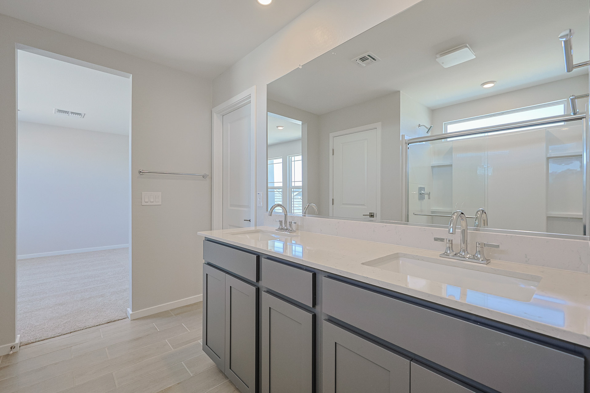 Modern bathroom with double sinks, gray cabinets, large mirror, and walk-in shower; view into carpeted adjacent room.