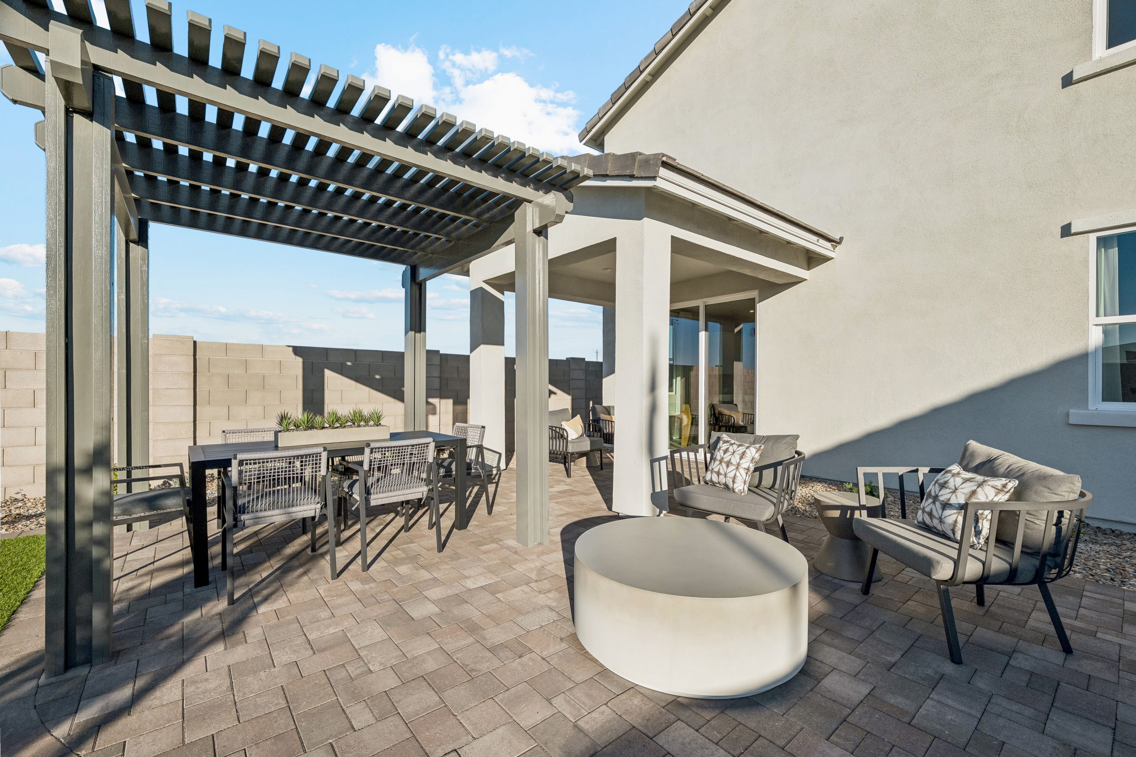 Modern outdoor patio with a pergola, dining table, chairs, lounge seating, and round coffee table on a paved surface adjacent to a house and block wall.