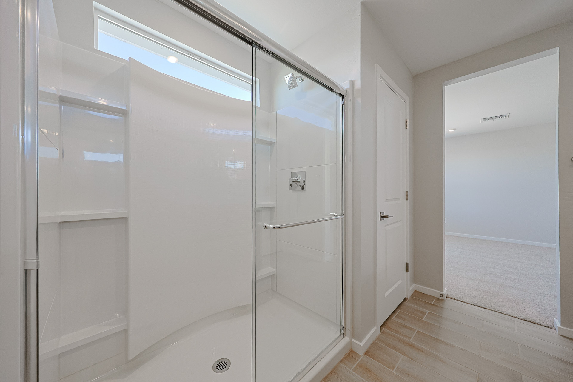 A modern bathroom with a glass-enclosed shower, built-in shelves, tile floor, and a white door leading to a carpeted room.