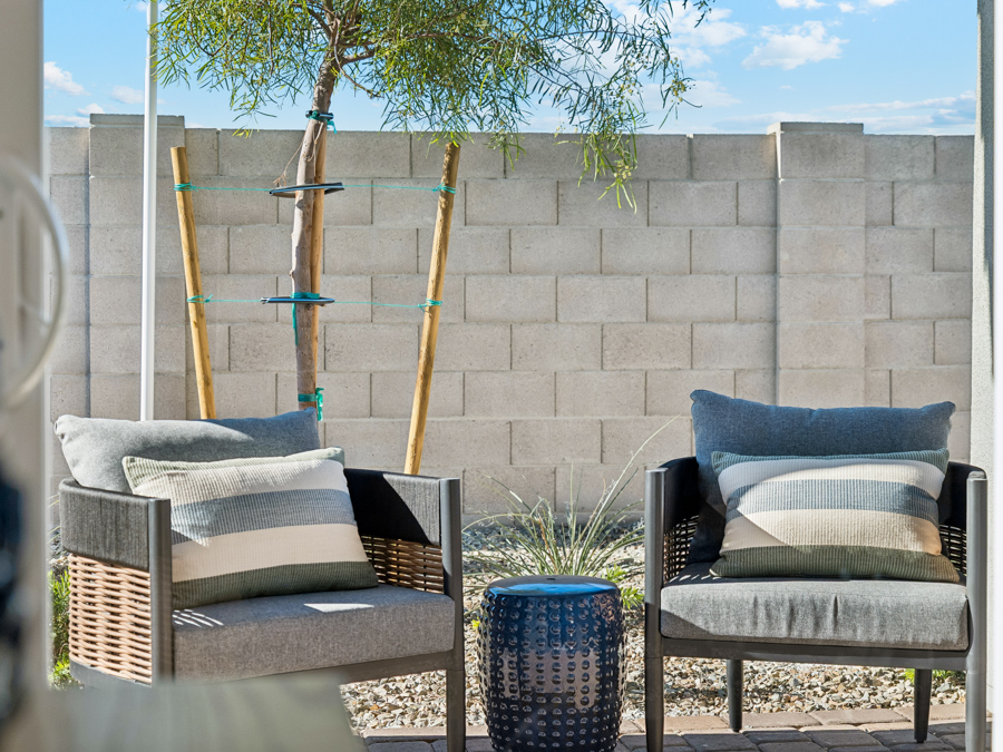 Two cushioned patio chairs with striped pillows sit beside a small blue table on a gravel yard, with a young tree and a concrete block wall in the background.