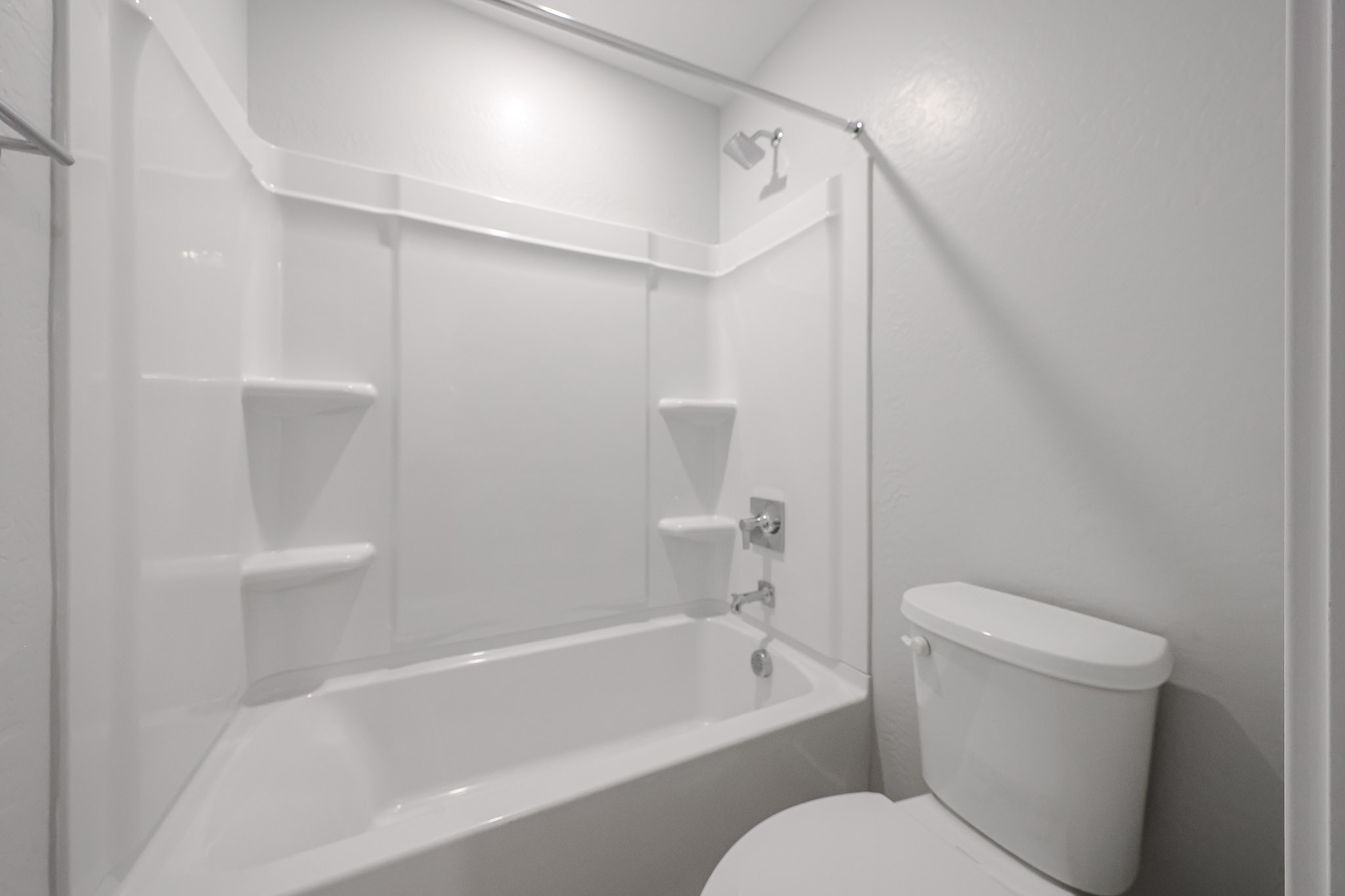 White bathroom with a bathtub-shower combo, built-in shelves, a curtain rod, and a toilet against light gray walls. The space appears clean and minimal.