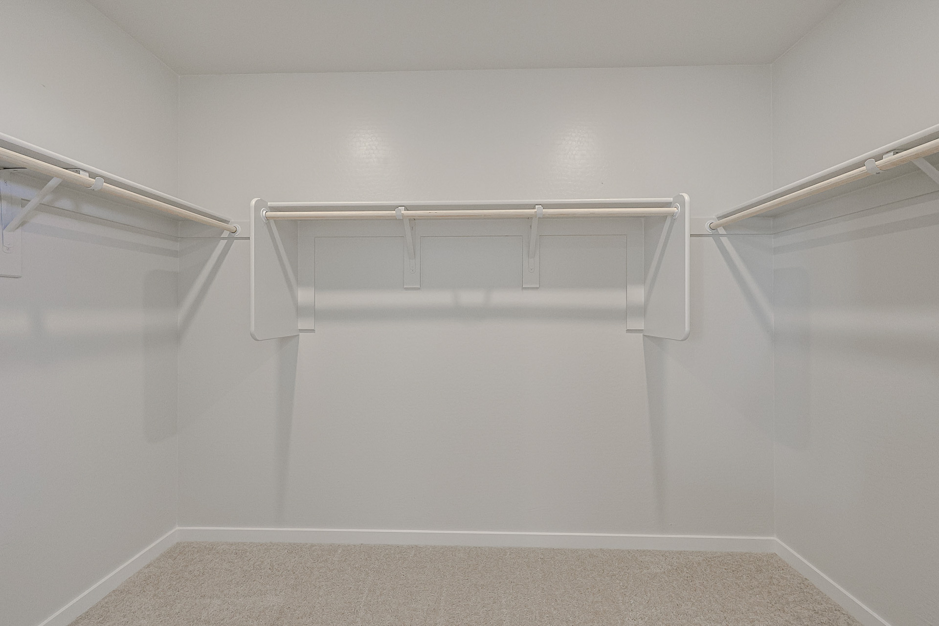 Empty walk-in closet with beige carpet, white walls, and three white hanging rods on adjacent walls.