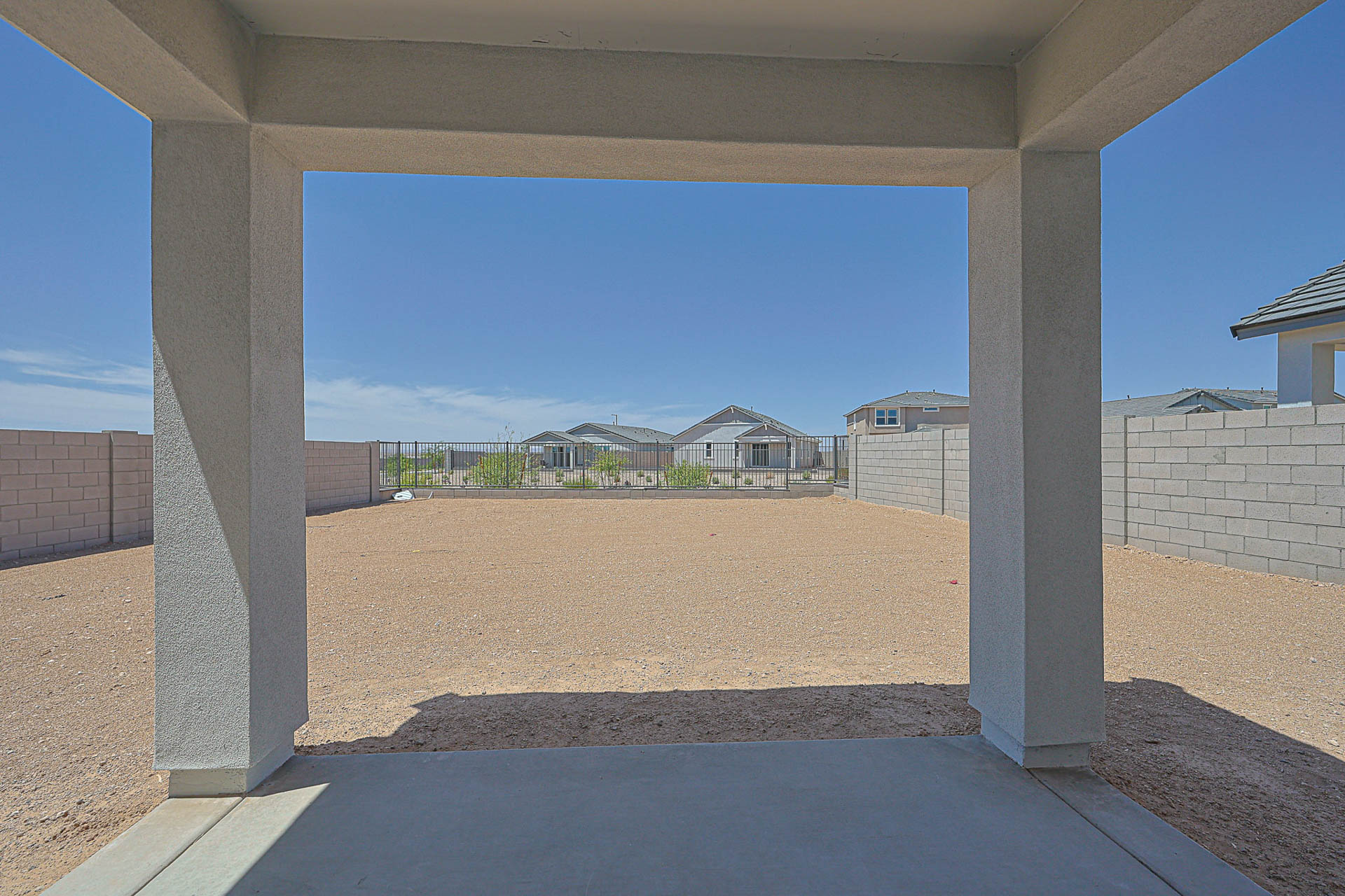 View from a covered patio looking out onto a large, empty, fenced backyard with gravel ground, under a clear blue sky.