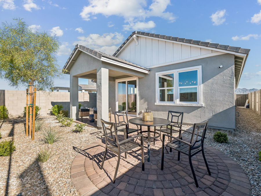 Modern backyard with a round patio, metal outdoor dining set, gravel landscaping, young tree, and a single-story gray house under a blue sky with clouds.